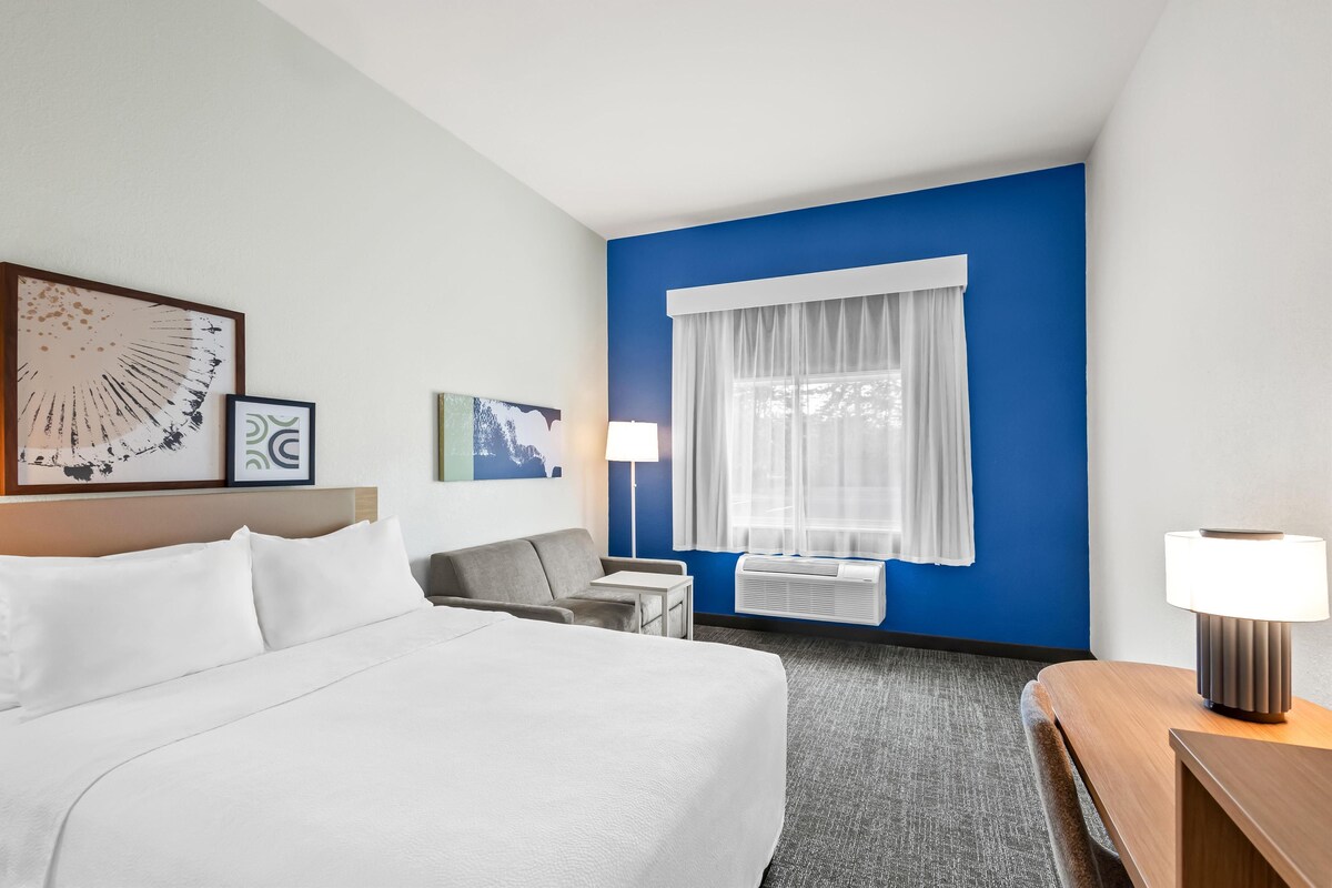 A hotel room with a large bed, a desk, a chair, a lamp, and artwork on the wall.