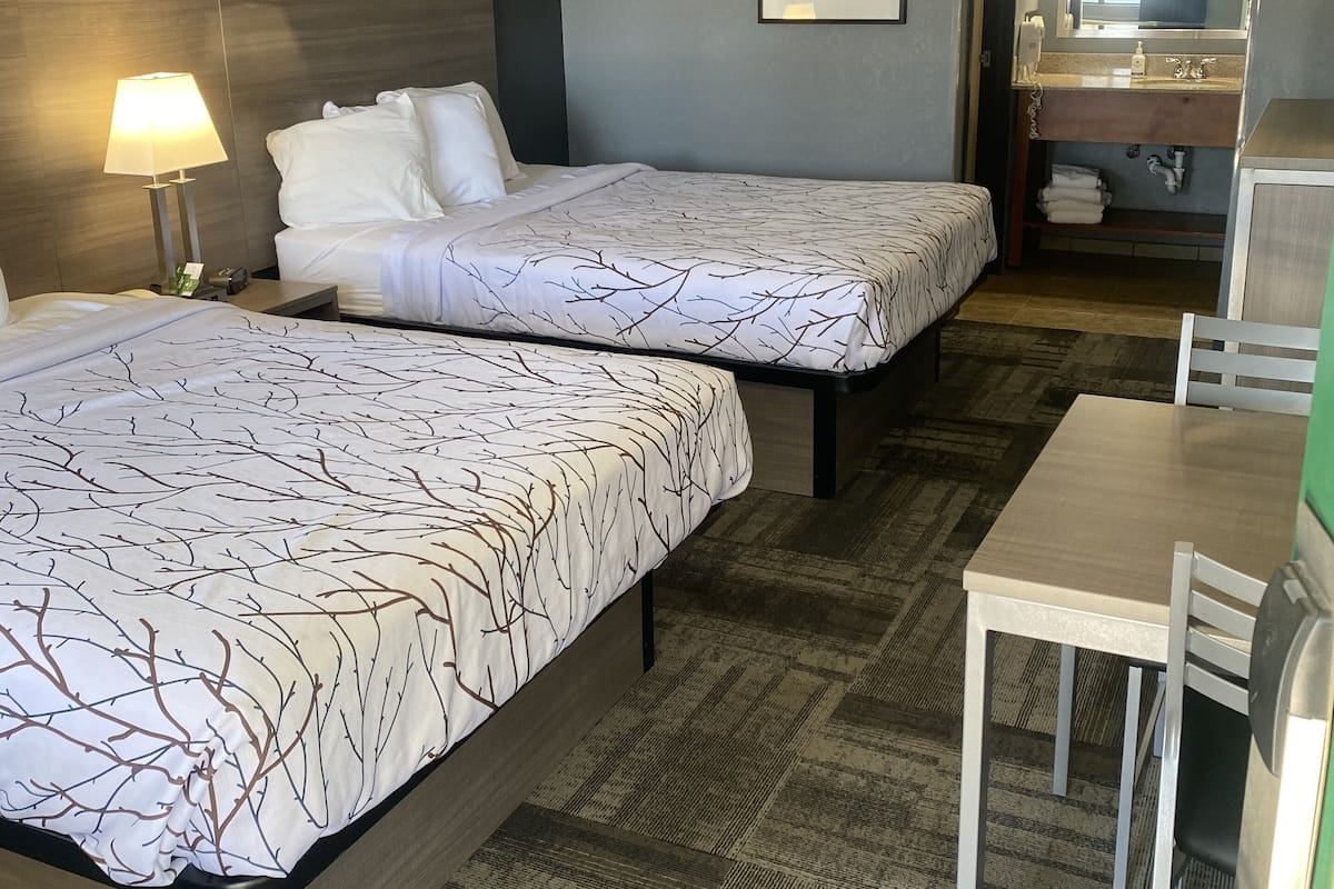 A hotel room with two beds, a desk, a chair, a lamp, a framed picture on the wall, and a bathroom with a mirror and sink.