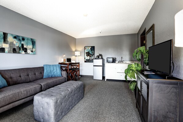 A compact living space with a grey sofa, ottoman, kitchenette, and a flat-screen TV on a cabinet. - GreenTree Inn of Prescott Valley (Prescott Valley)
