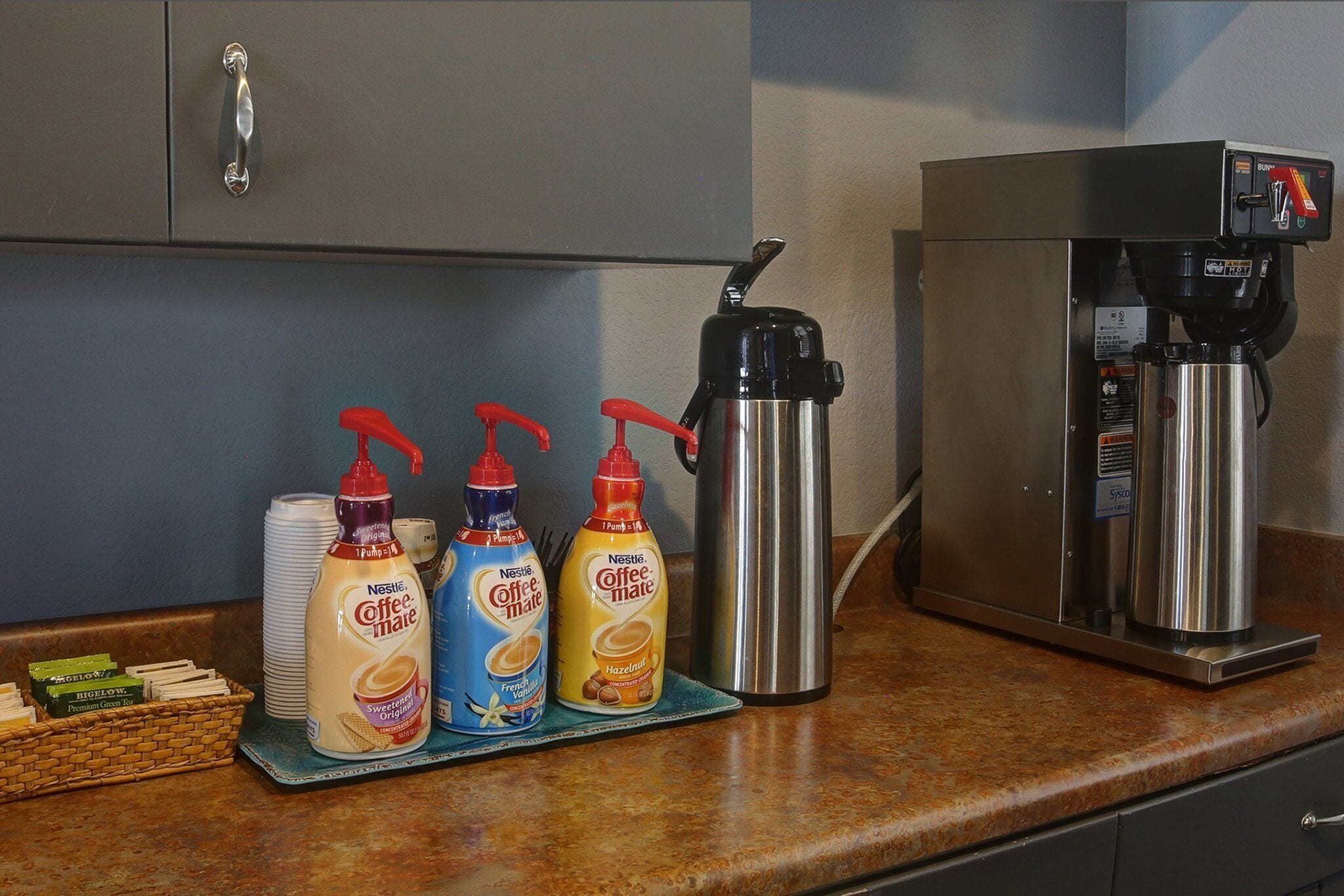 A kitchen area with a coffee machine, three bottles of coffee creamer, and a stainless steel thermos.
