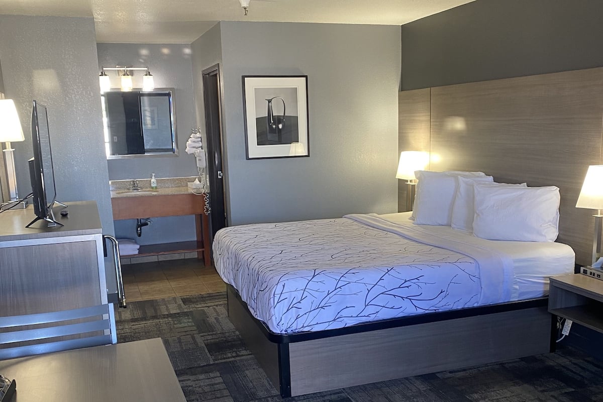 A hotel room with a bed, bedside tables, a desk, a chair, a bathroom with a sink and mirror, and a ceiling fan.