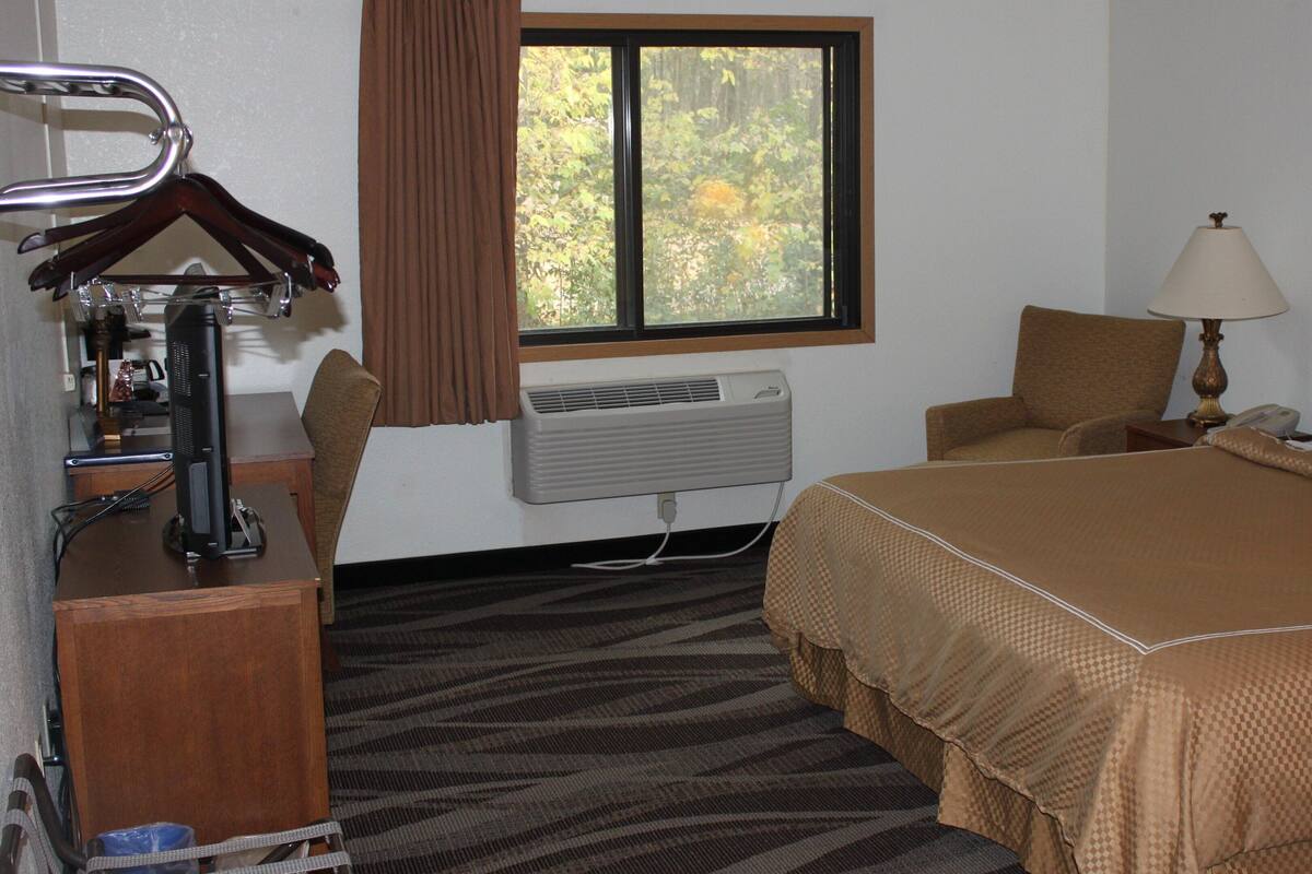A hotel room with a bed, a chair, a desk with a computer, a lamp, and an air conditioner.