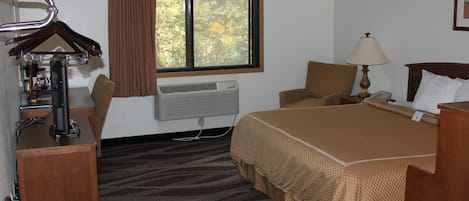 A hotel room with a bed, a chair, a desk with a computer, a lamp, and an air conditioner.