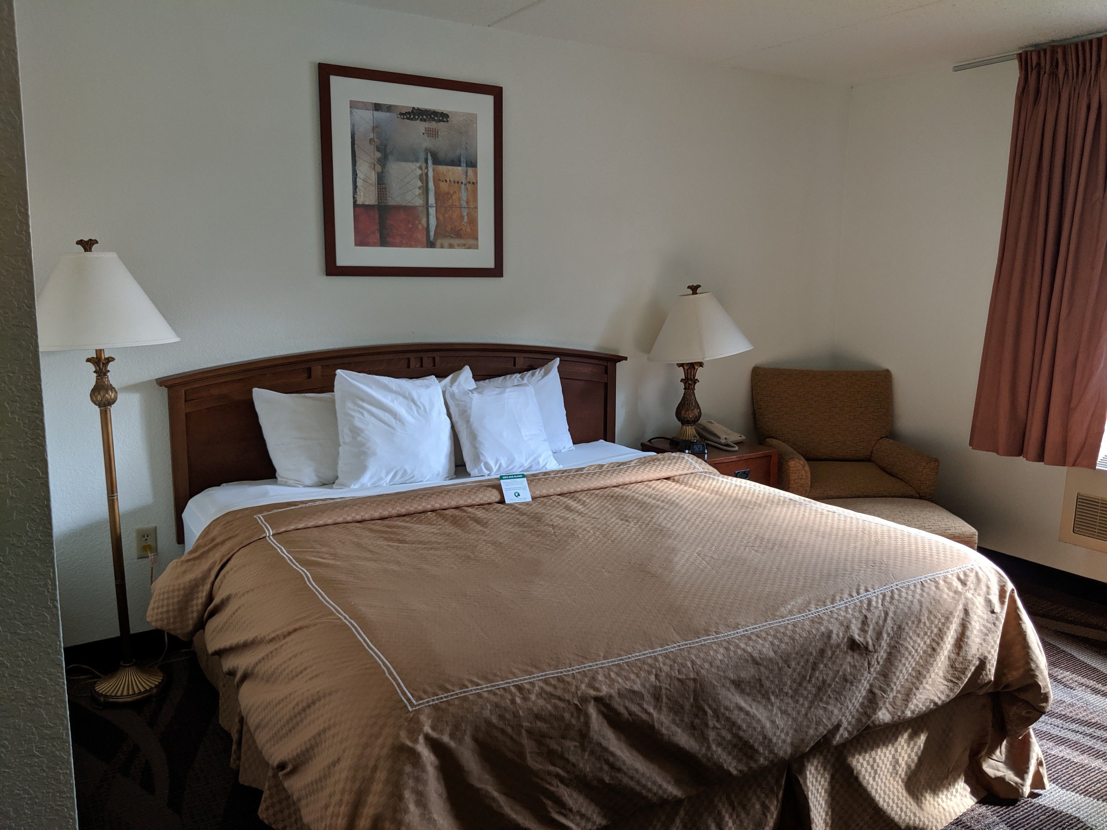 A hotel room with a large bed, two bedside lamps, a chair, a framed picture on the wall, and a window with curtains.