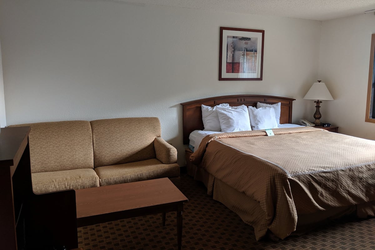A hotel room with a bed, a sofa, a small table, a nightstand with a lamp, and a framed picture on the wall.