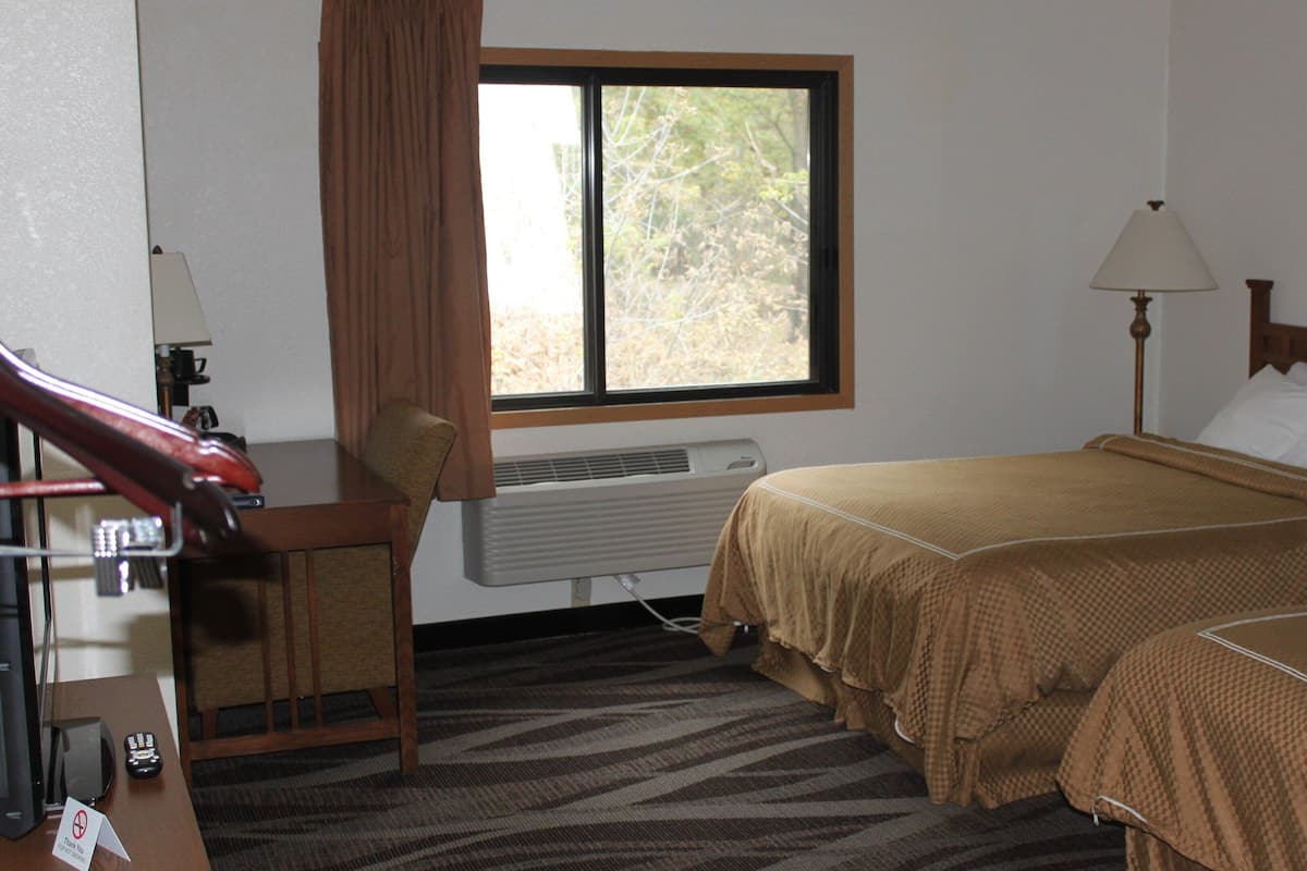 A hotel room with a desk, chair, two beds, a window, a lamp, and a framed picture on the wall.