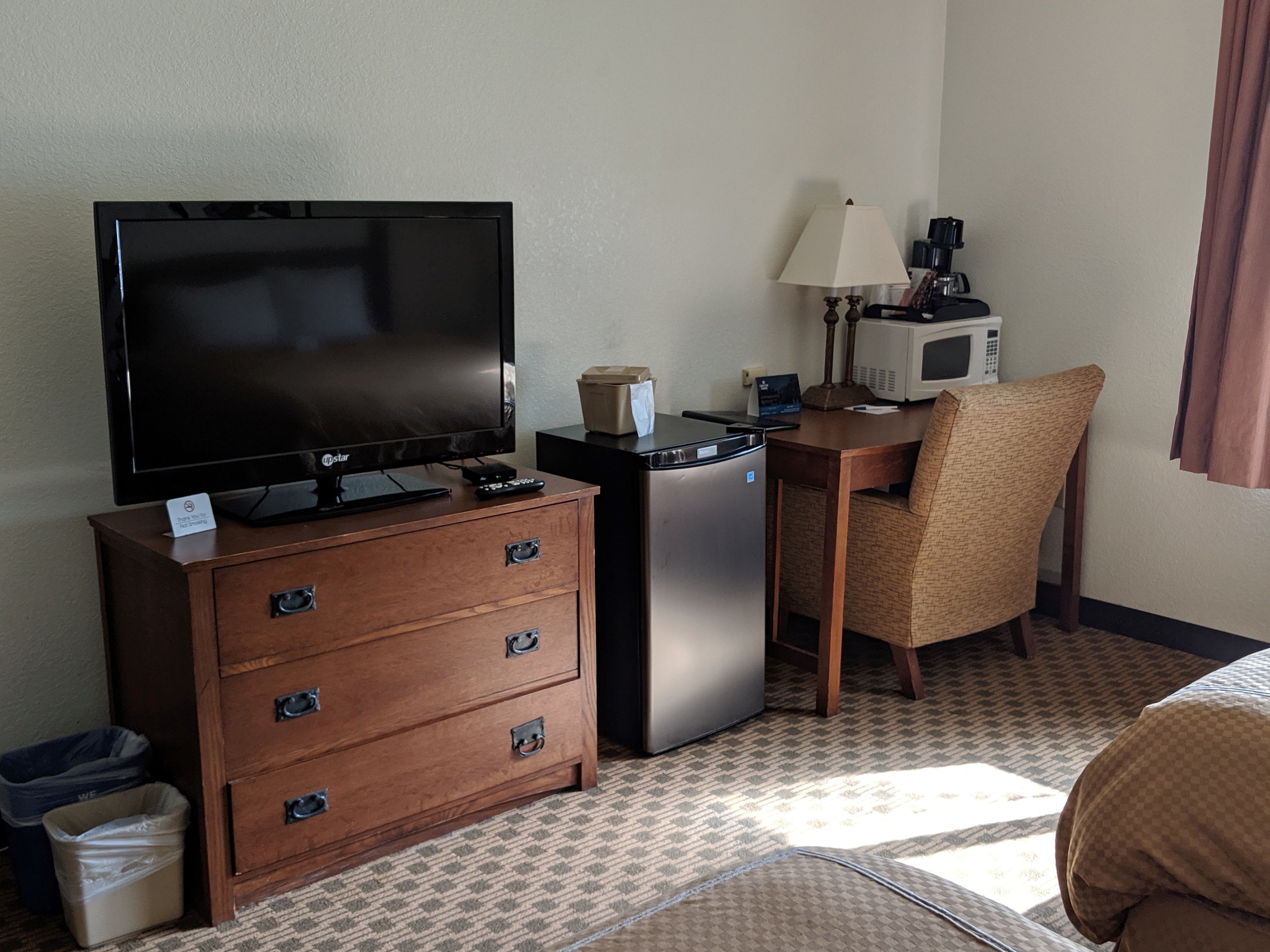 Habitación de hotel con televisión de pantalla plana, cómoda de madera, refrigerador, microondas, lámpara y una silla.