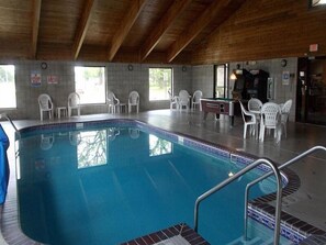 An indoor swimming pool with a tiled edge, surrounded by white plastic chairs, and a wooden ceiling.