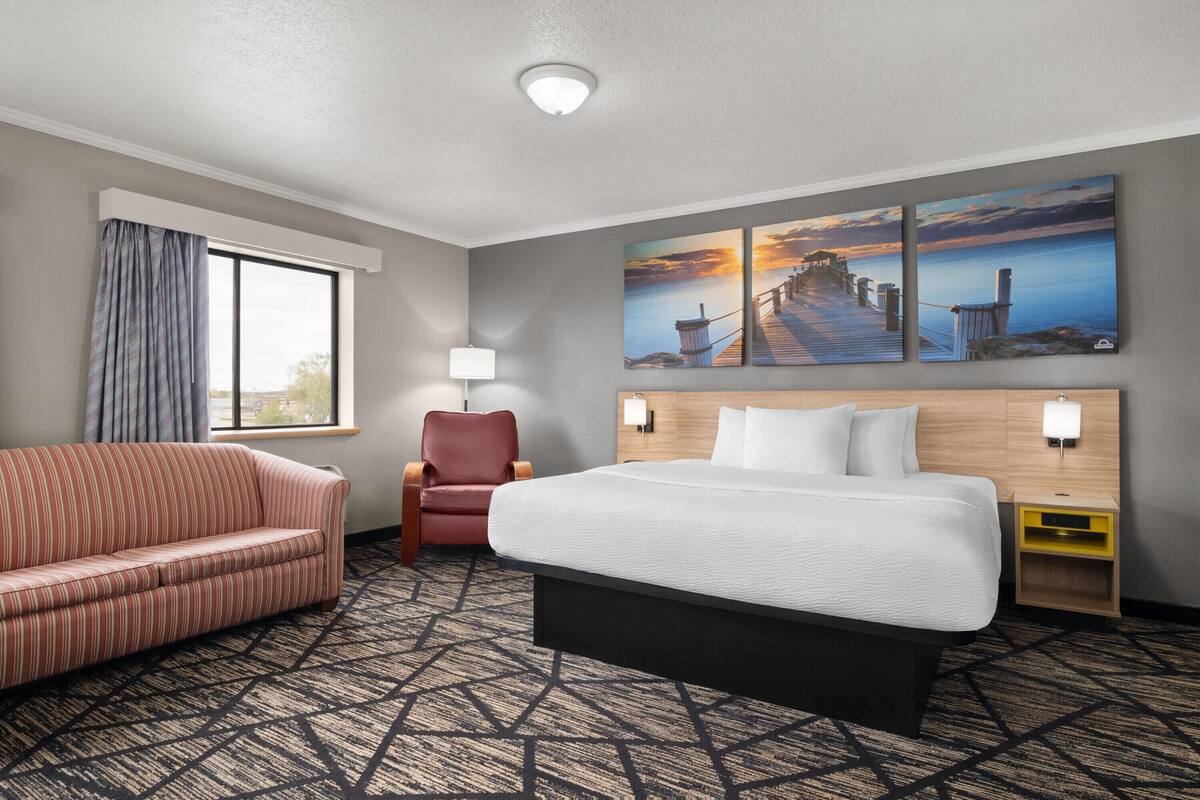 A hotel room with a bed, a sofa, a chair, a window with curtains, a wall-mounted lamp, and a painting of a pier.