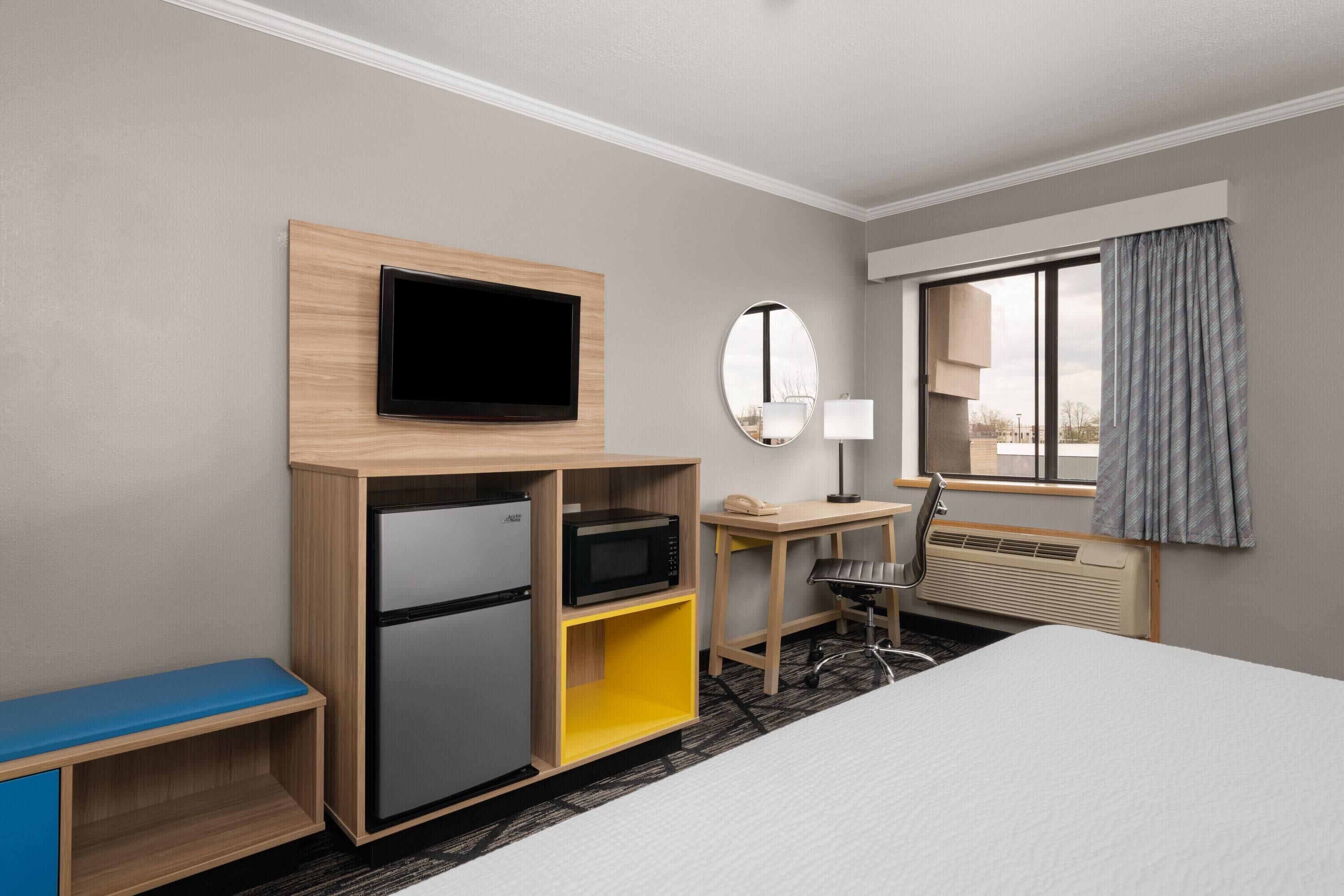 A hotel room with a bed, a desk, a television, a microwave, a refrigerator, a mirror, a window with curtains, and a chair.