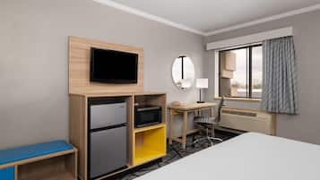 A hotel room with a bed, a desk, a television, a microwave, a refrigerator, a mirror, a window with curtains, and a chair.