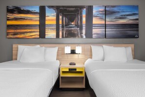 A hotel room with two beds, a nightstand with a clock, and a large sunset painting above the beds. - La Quinta Inn by Wyndham Milwaukee Airport / Oak Creek (Oak Creek)