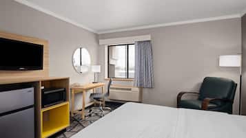 Habitación de hotel con cama, escritorio con silla, televisión, microondas, refrigerador, ventana con cortinas y lámpara.