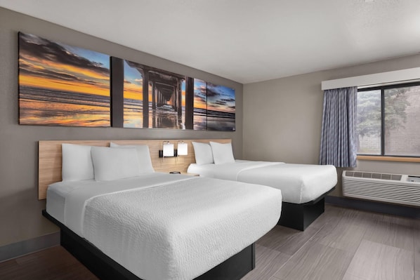A hotel room with two beds, a large window, a wall-mounted air conditioner, and three paintings.