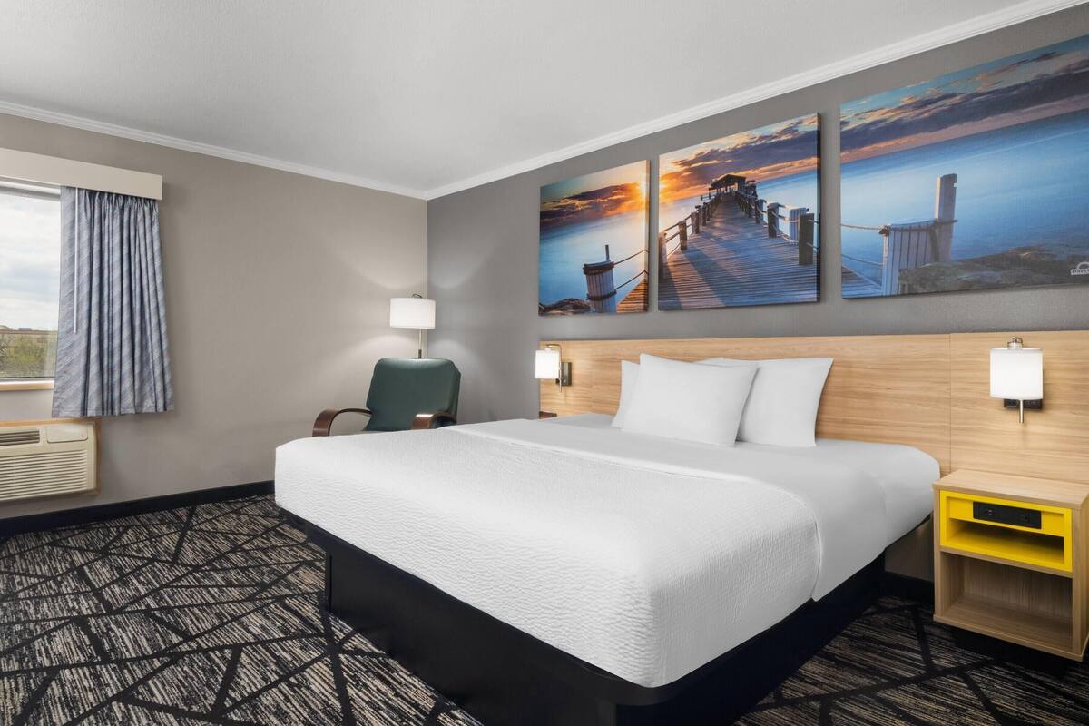 A hotel room with a large bed, a desk, a chair, a lamp, and two paintings on the wall.