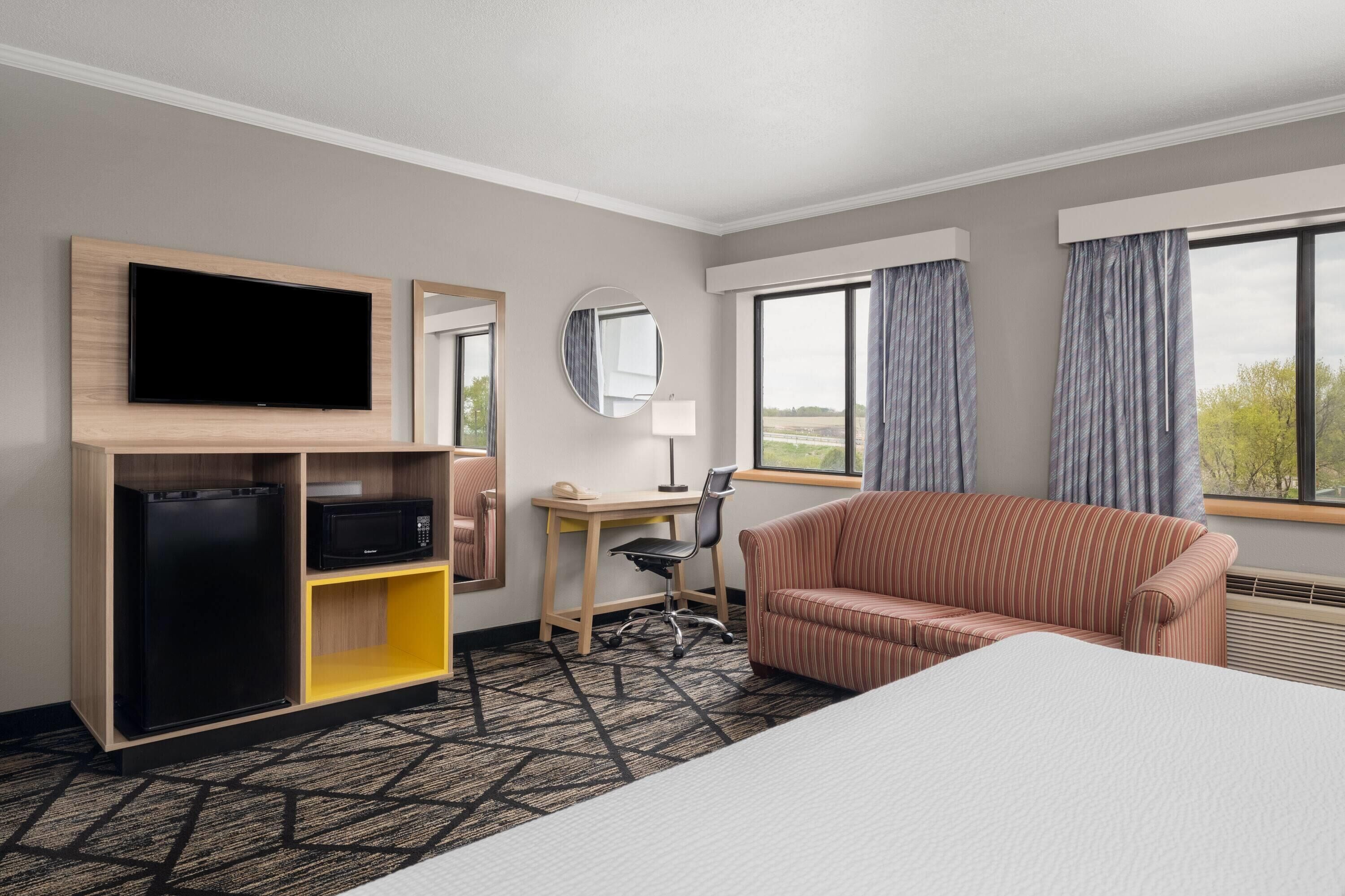 A hotel room with a bed, a TV, a desk, a chair, a sofa, a mirror, and a window with curtains.