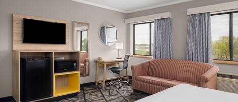 Habitación de hotel con cama, televisor, escritorio, silla, sofá, espejo y ventana con cortinas.