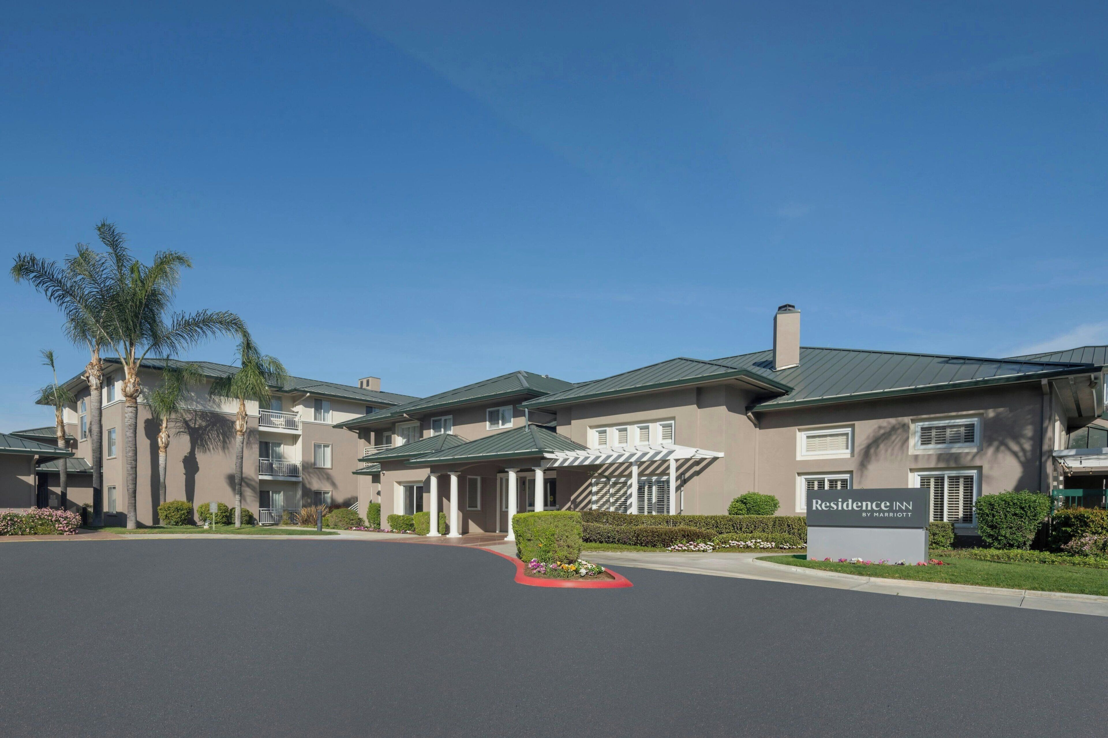 A multi-story hotel with a sign reading 'Residence Inn by Marriott,' featuring a central entrance with a covered walkway, palm trees, and well-maintained landscaping.