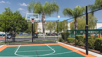 An outdoor basketball court with a net, surrounded by a fence, palm trees, and a road with cars in the background.