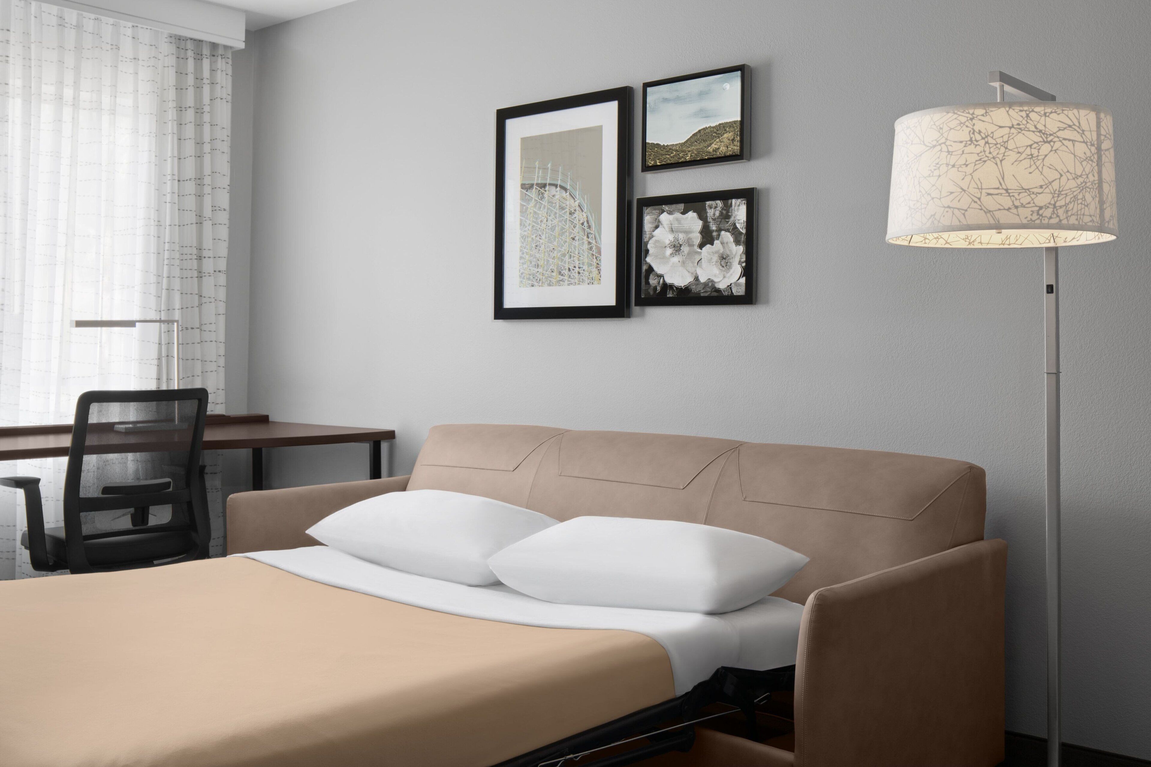 A hotel room with a bed, a desk, and a chair. There are two framed pictures on the wall.