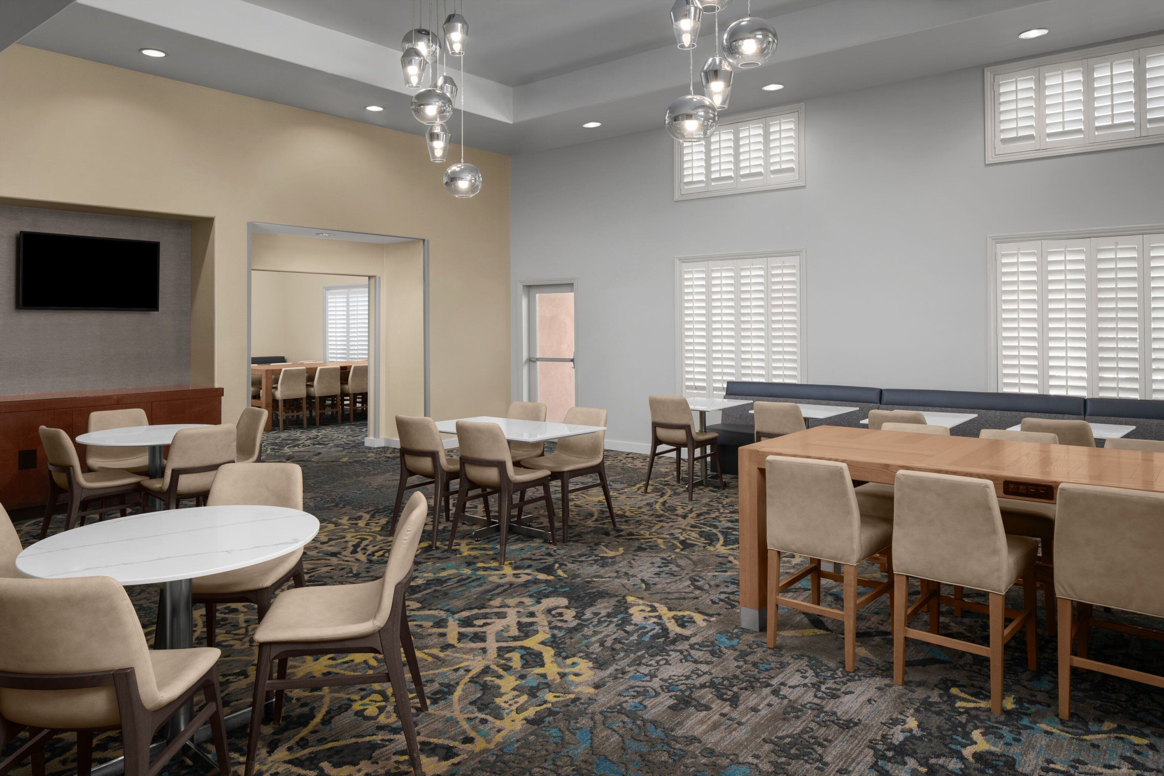 A conference room with multiple tables and chairs, a flat-screen TV mounted on the wall, and large windows with blinds.