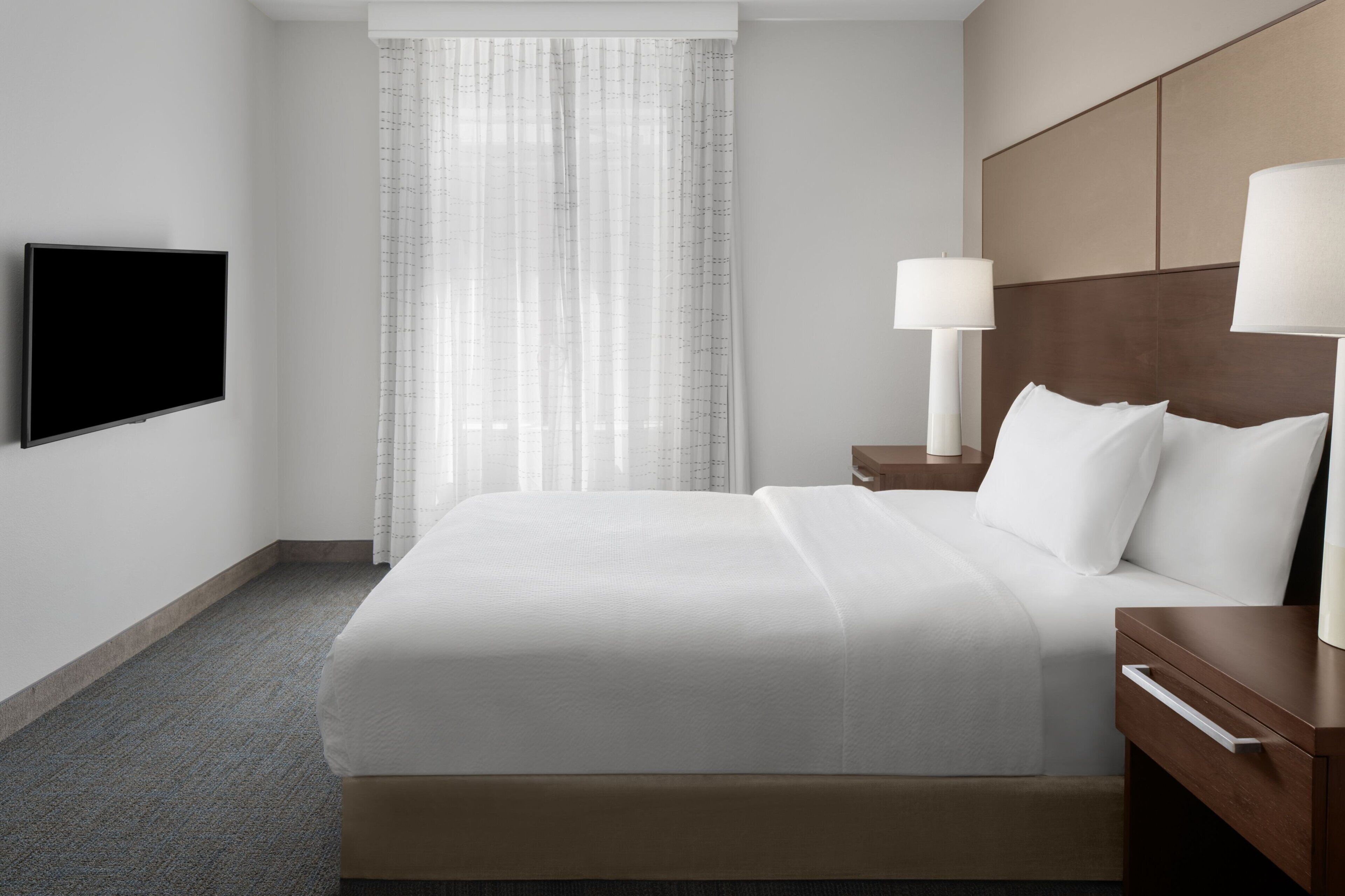 Habitación de hotel con una cama grande, un televisor de pantalla plana montado en la pared, una mesita de noche con lámpara y una ventana con cortinas traslúcidas.
