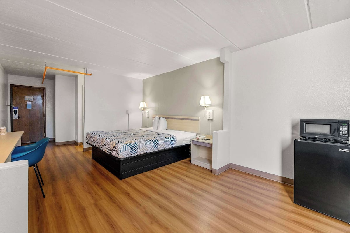 A hotel room with a bed, bedside lamps, a desk, a chair, a microwave, and a refrigerator.