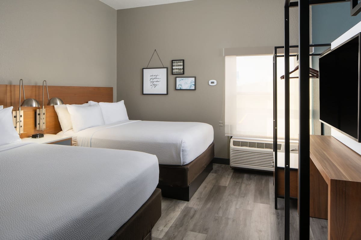 A hotel room with two beds, a wooden headboard, a flat-screen TV, a wall-mounted air conditioner, and framed pictures on the wall.