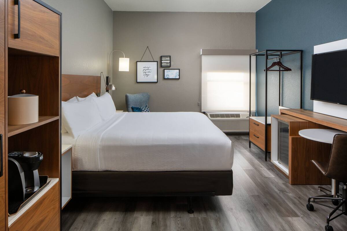 A modern hotel room with a large bed, a desk with a chair, a TV, a coffee machine, and a wardrobe.