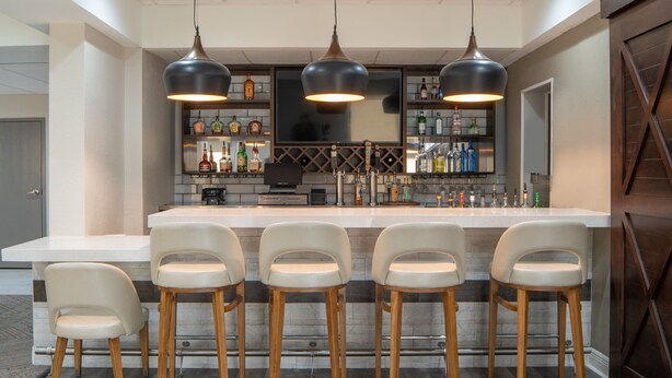Uma área de bar com um balcão comprido, cinco banquetas e uma variedade de garrafas nas prateleiras.