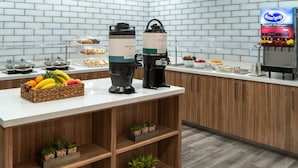Balcão de pequeno-almoço com máquinas de café, uma cesta de fruta e dispensador de sumo.