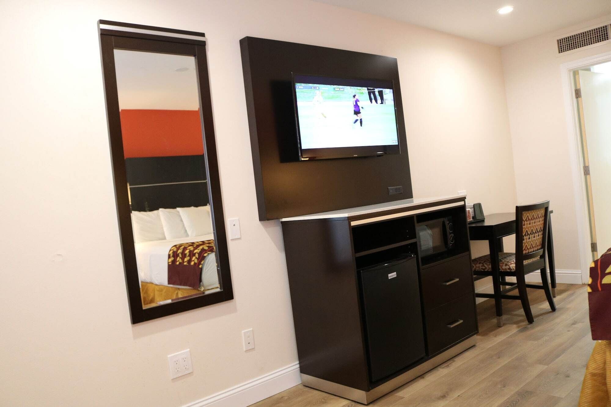 Un espacio habitable compacto con un televisor de pantalla plana montado sobre una kitchenette, una zona de comedor con mesa y sillas, y un espejo grande en la pared.