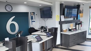 A modern reception area with a large number '6' on the wall, a digital clock, a flat-screen TV, and a coffee station. - Motel 6 Vallejo, CA – Napa Valley (Vallejo)