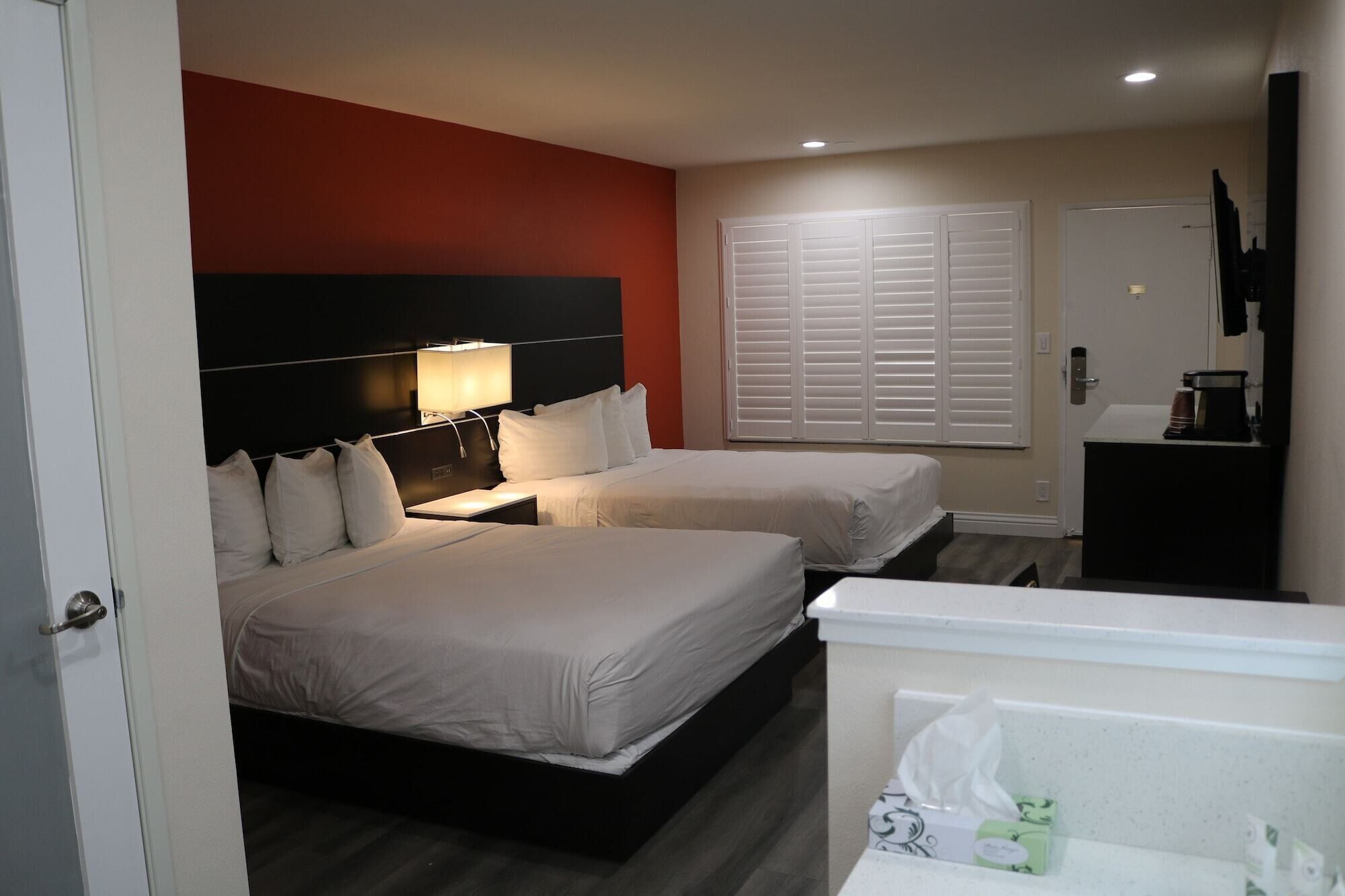 Quarto de hotel com duas camas, uma mesa de cabeceira com luminária, uma janela com persianas, uma pequena escrivaninha com cafeteira e um banheiro com dispensador de toalhas.