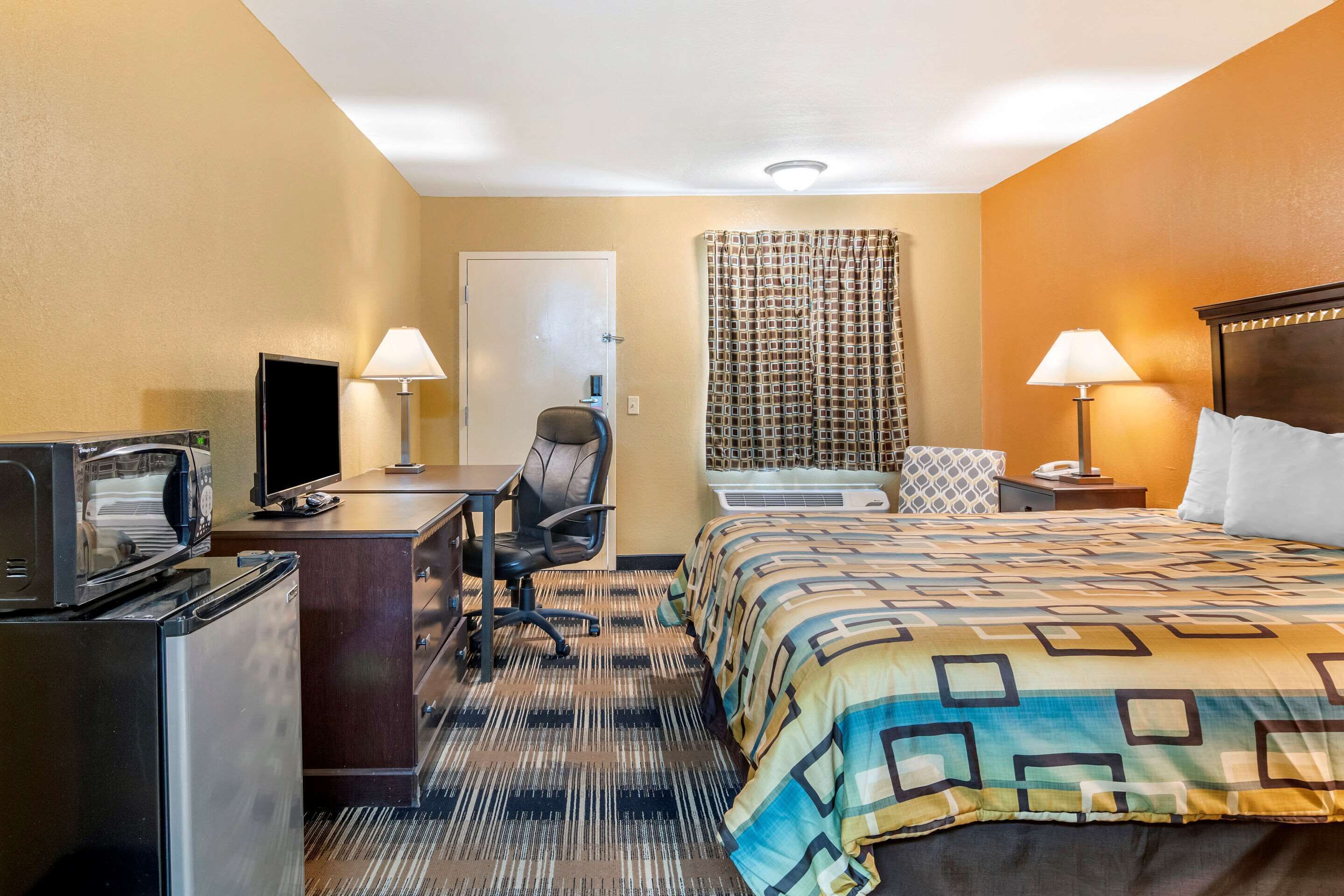 Habitación de hotel con cama, escritorio, silla, televisor, refrigerador y ventana con cortinas.