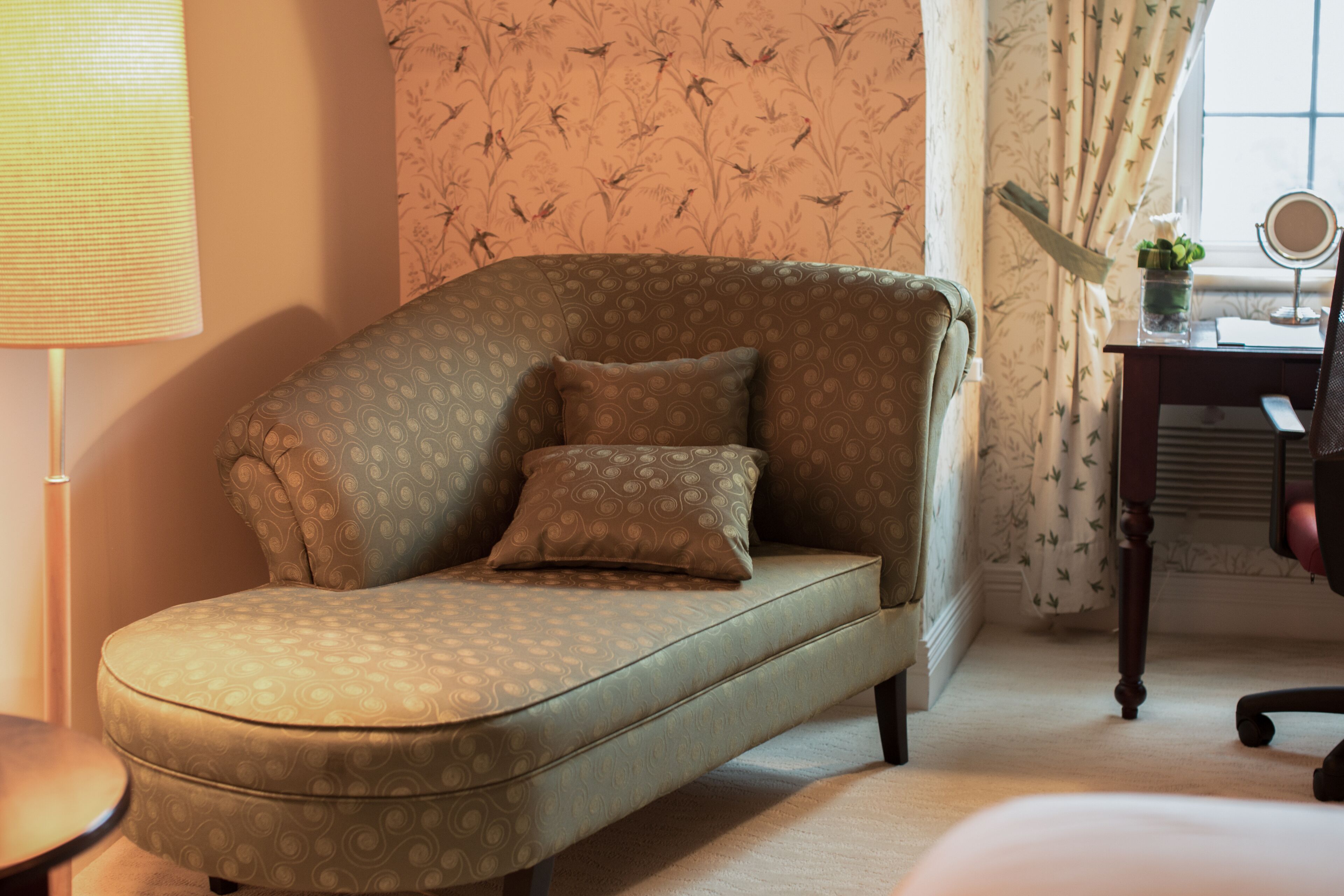 A room with a curved sofa, a small table, a lamp, a mirror, and a window with curtains.