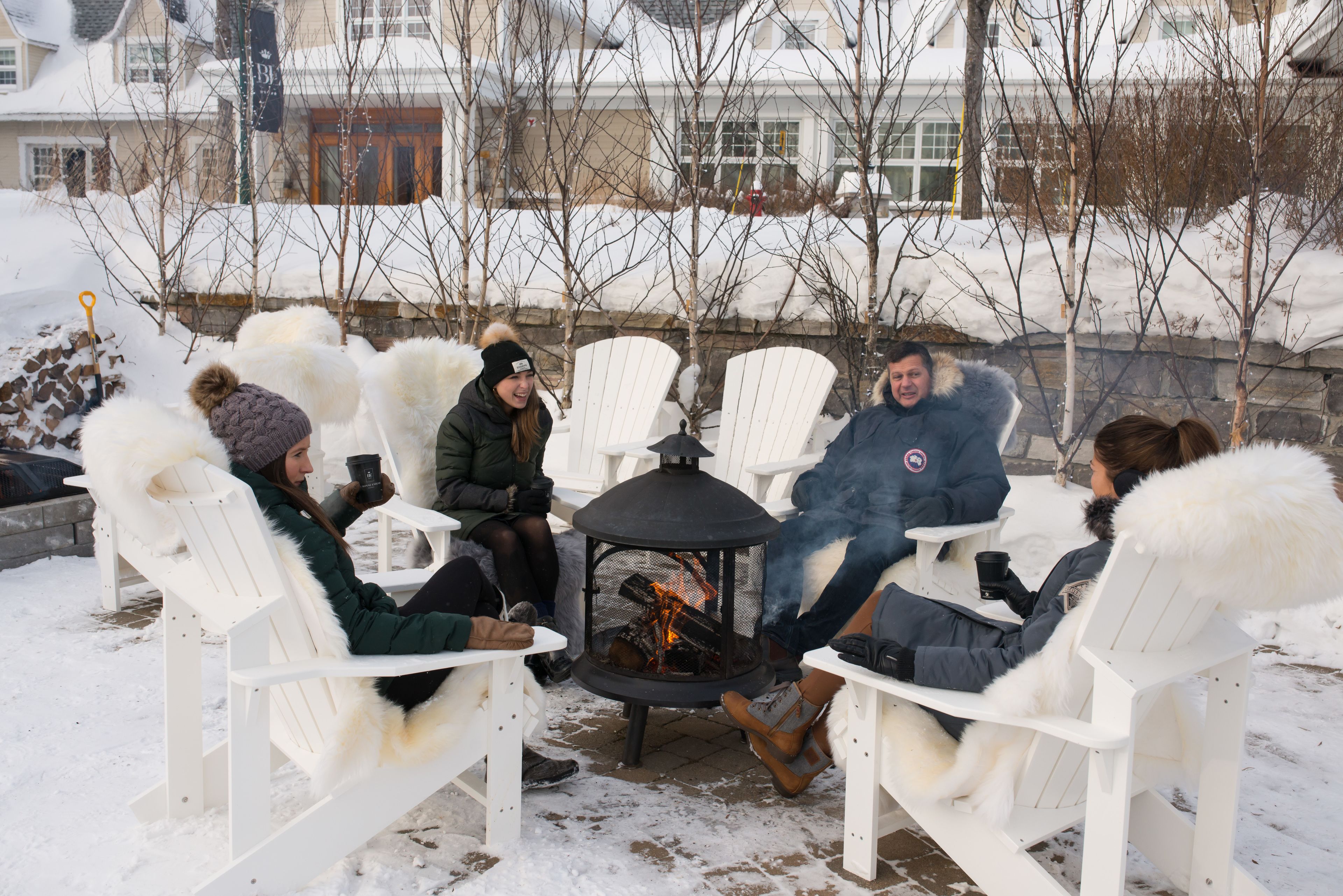 A group of people seated around a fire pit in white chairs with fur trim, in a snowy outdoor setting.