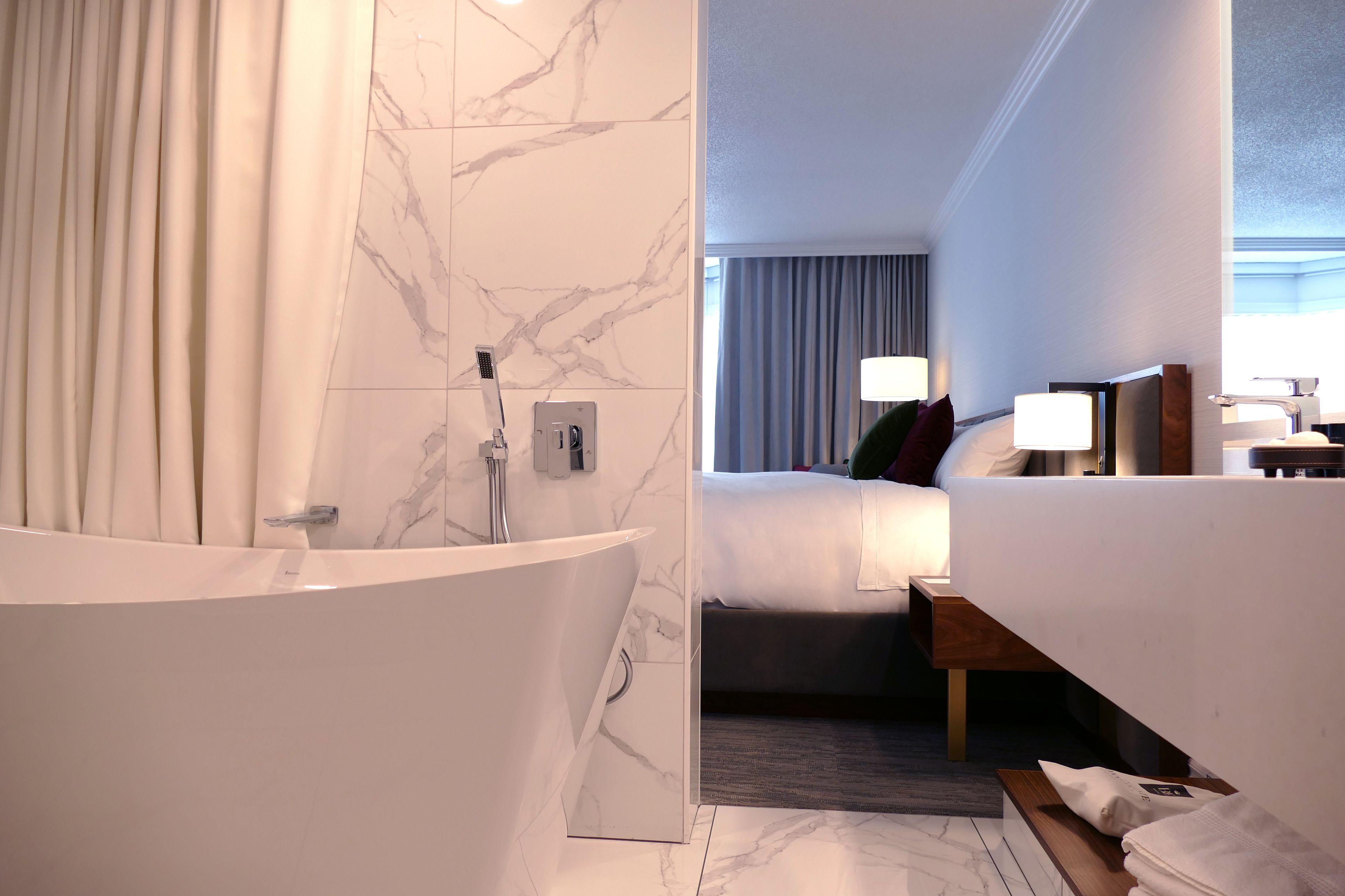 A modern hotel bathroom with a marble-tiled wall, a freestanding bathtub, and a visible bedroom with a bed, bedside table, and lamp.