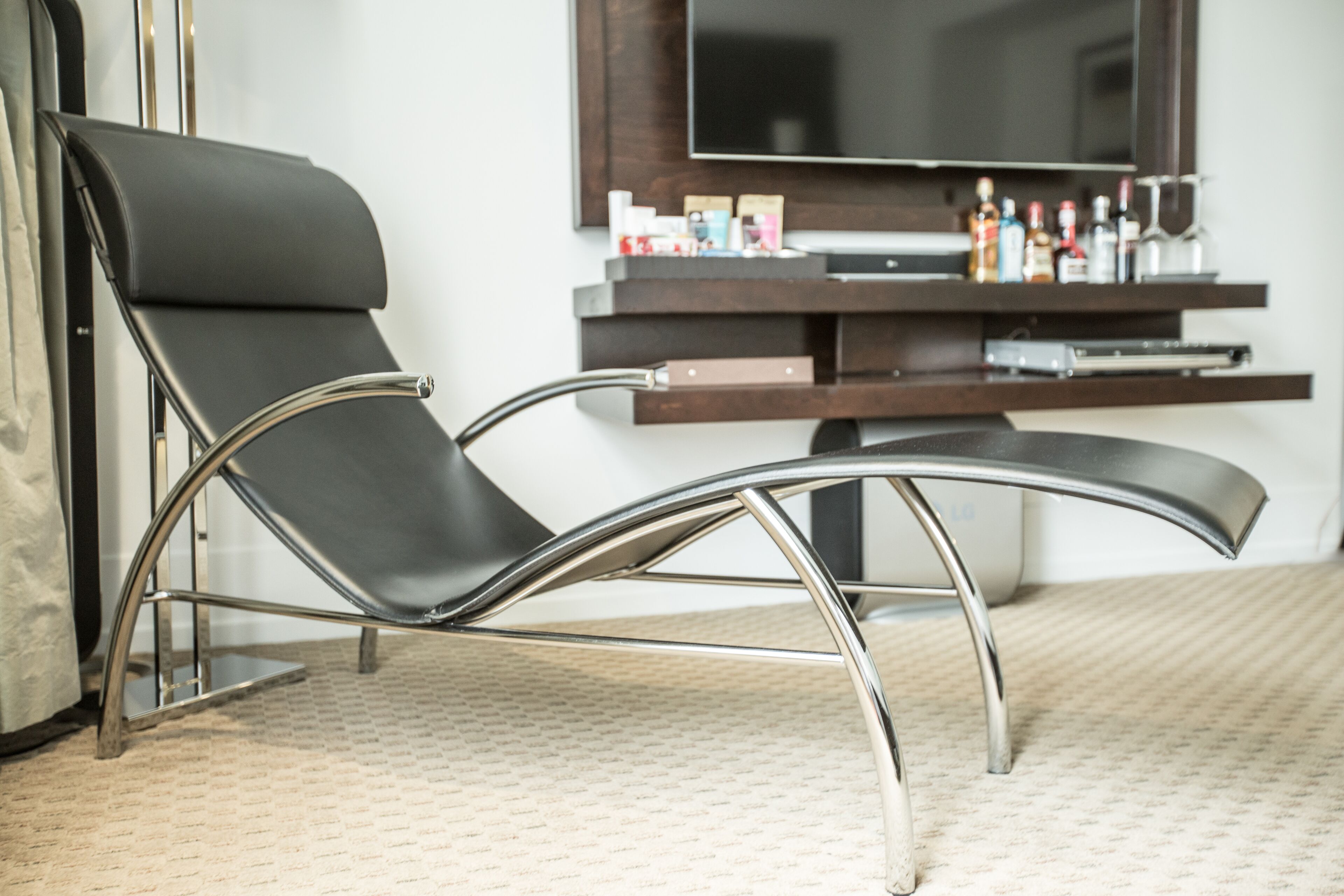 Una silla moderna con estructura metálica elegante y tapicería negra, ubicada en una habitación con un televisor de pantalla plana grande montado en la pared y un mueble bajo de madera con varios objetos.