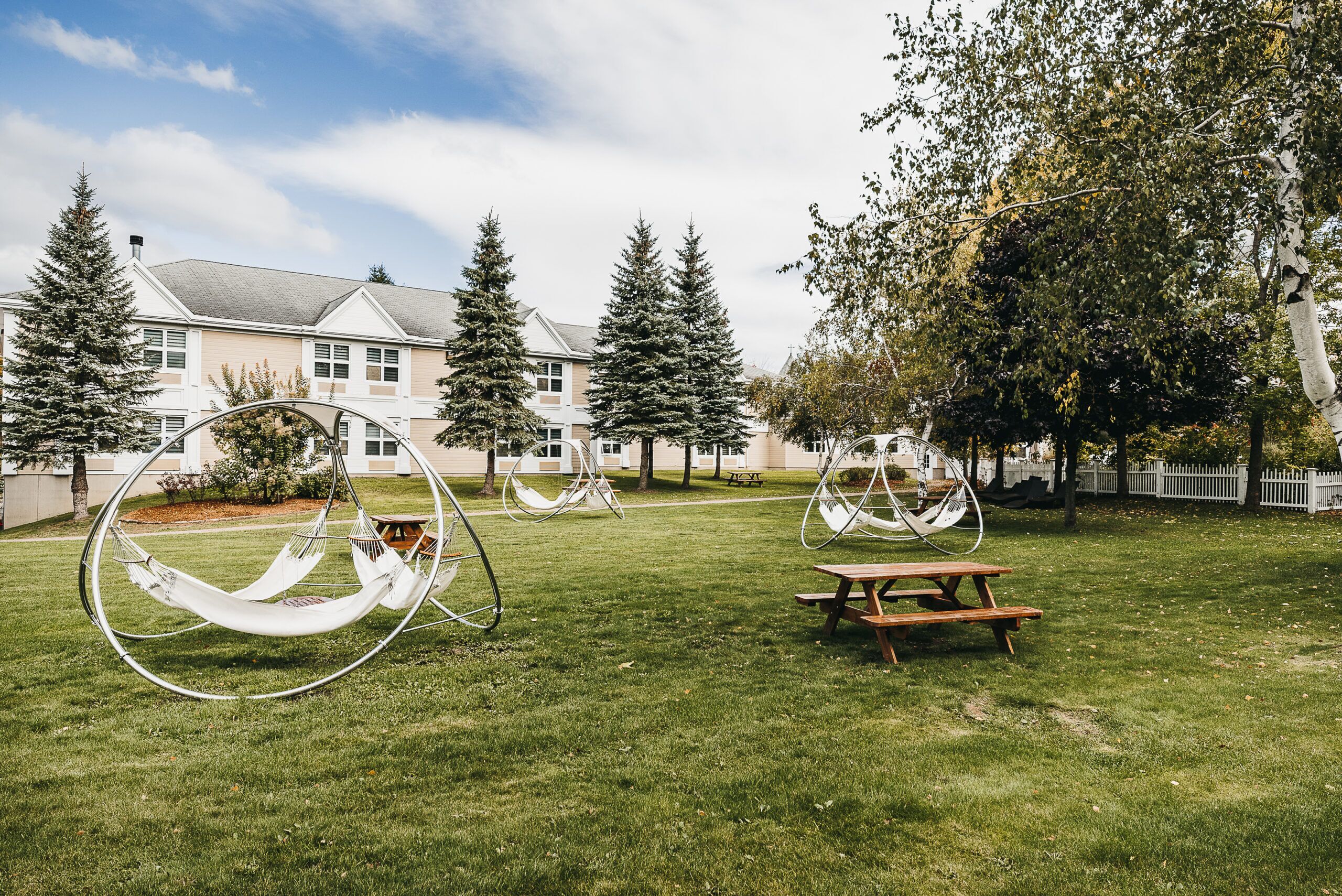 A grassy area with hammocks and a picnic table, surrounded by trees and a building with a white facade.
