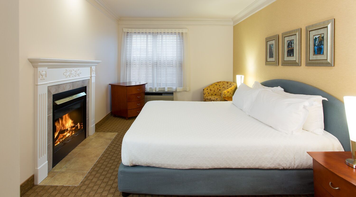 A hotel room with a fireplace, a bed, a chair, and framed pictures on the wall.