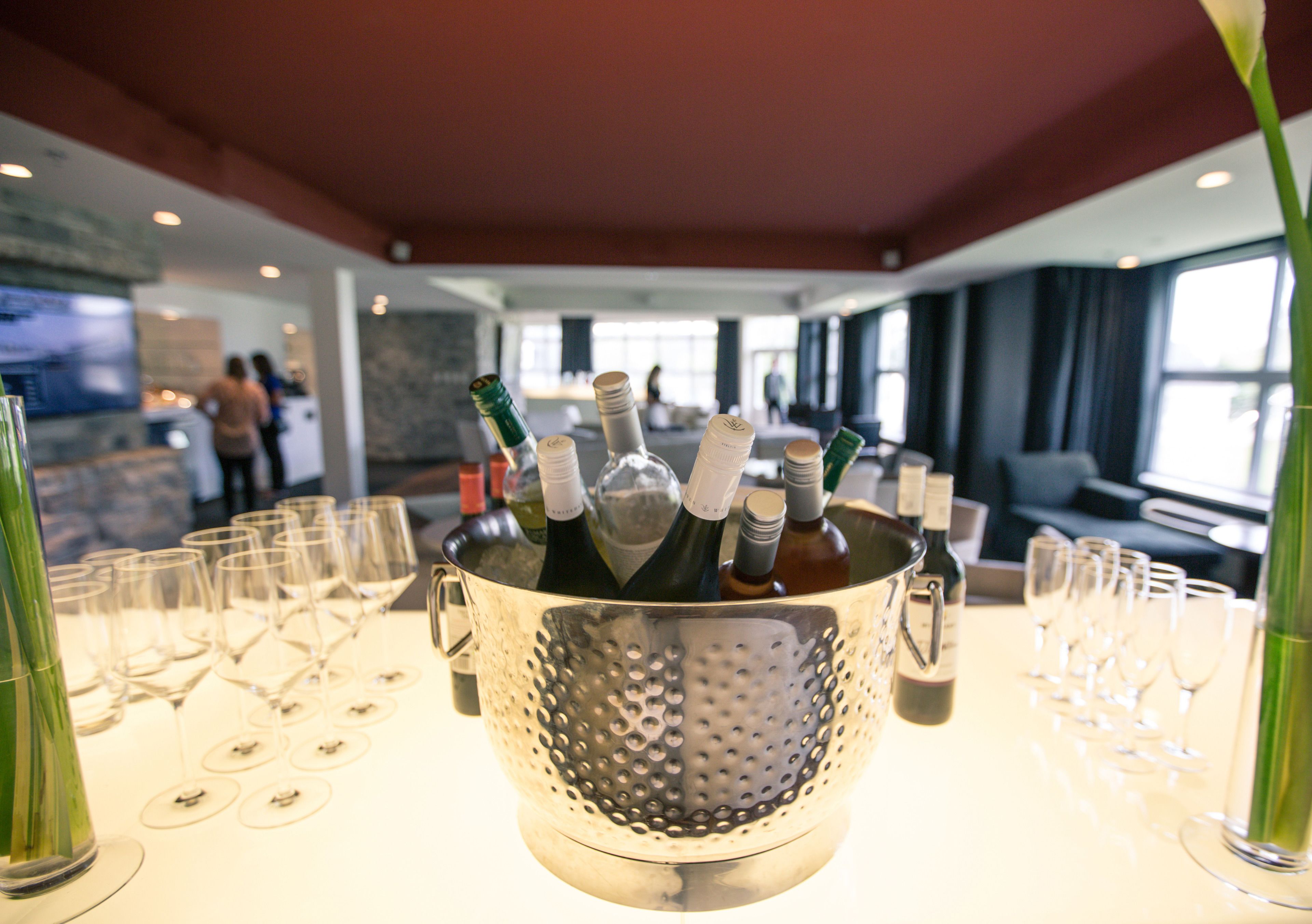 A metal container with wine bottles and glasses on a table in a room with large windows and a red ceiling.
