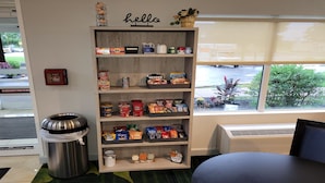 A snack shelf with various food items, a trash bin, a 'hello' sign, and a window with a view of the outside. - City Express By Marriott Shepherdsville Louisville (Louisville)