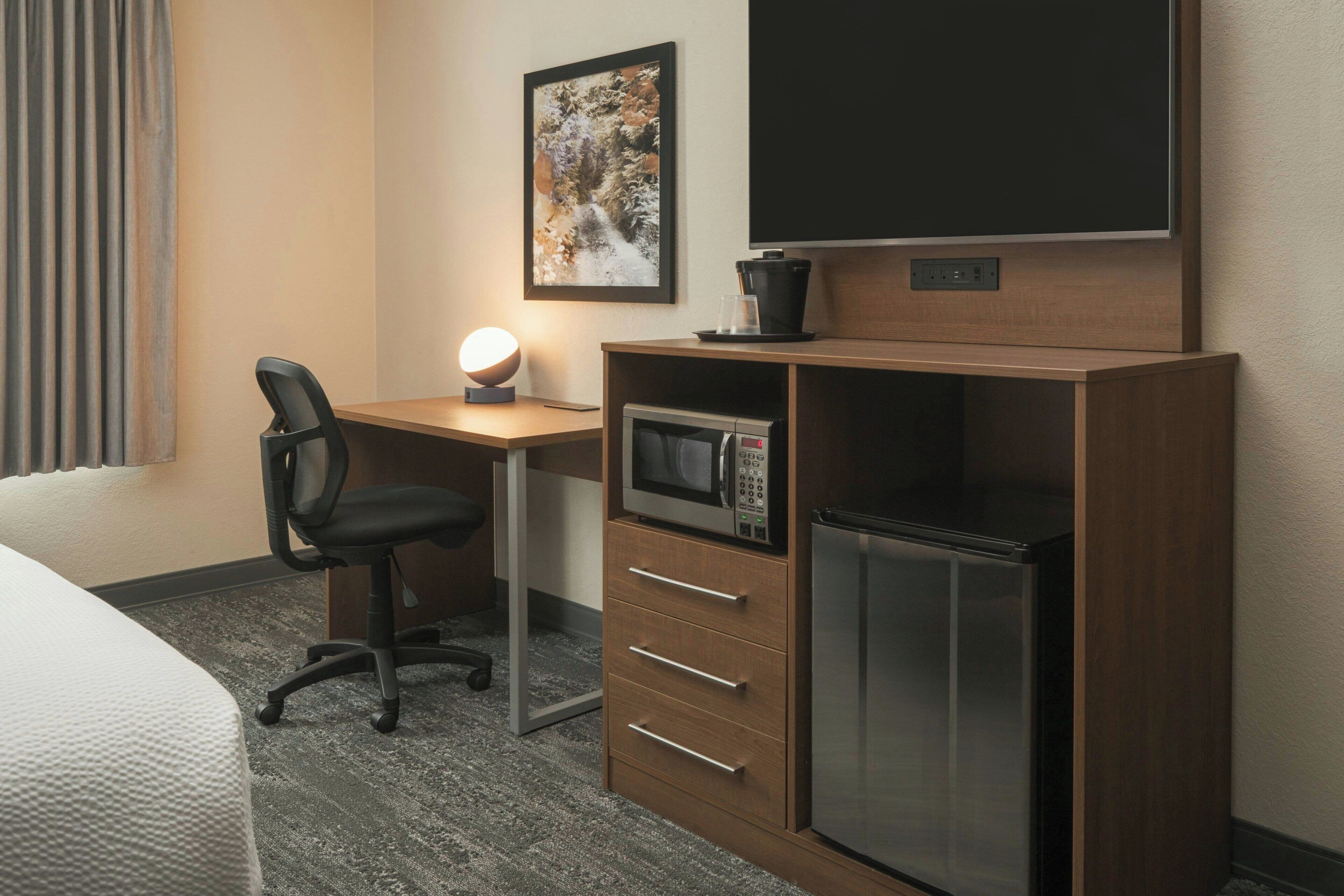 Habitación de hotel con cama, escritorio con silla, televisor de pantalla plana, microondas, refrigerador y cafetera.