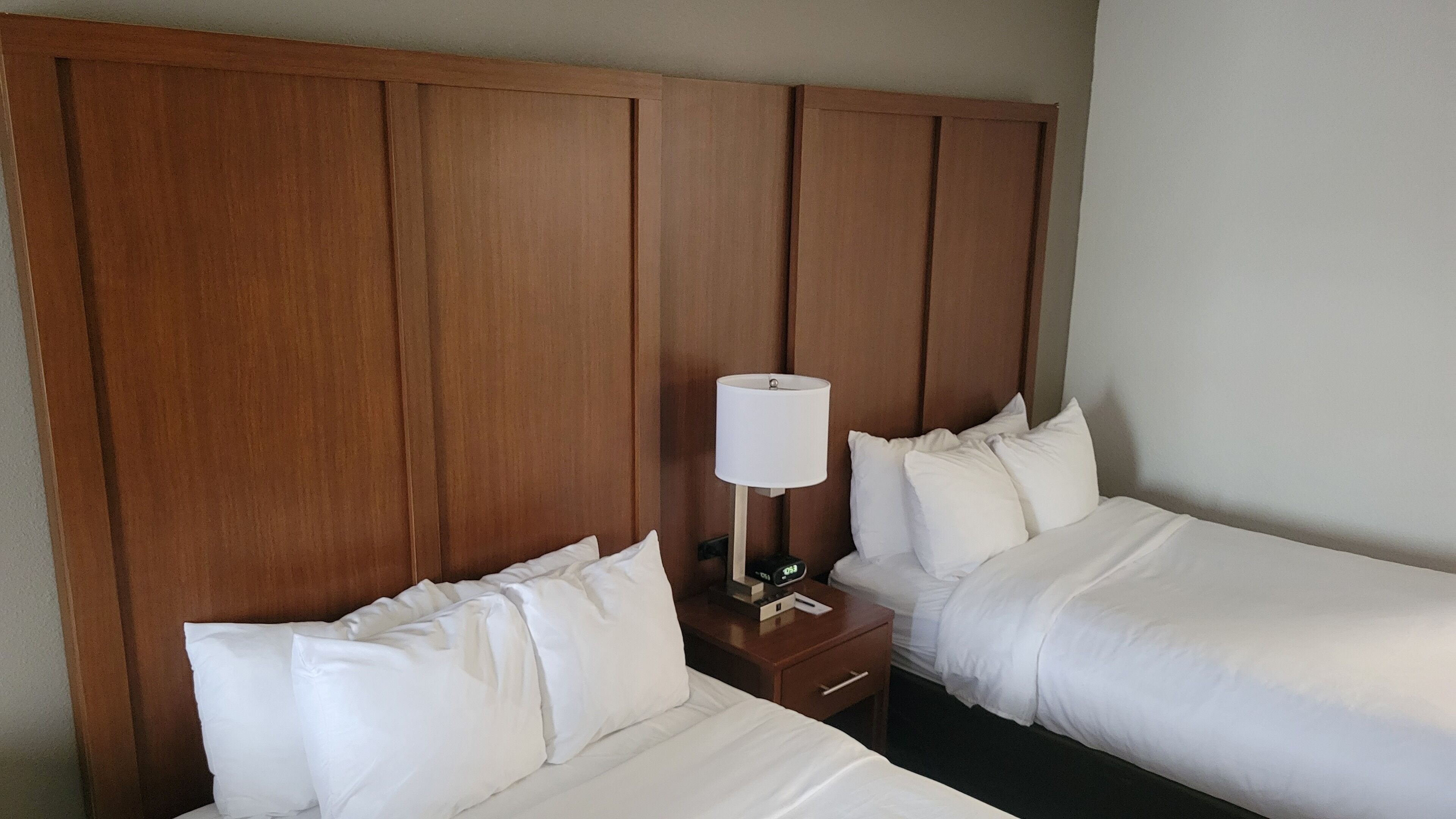 A hotel room with two single beds, wooden headboards, white bedding, a bedside table with a lamp, and a digital clock.