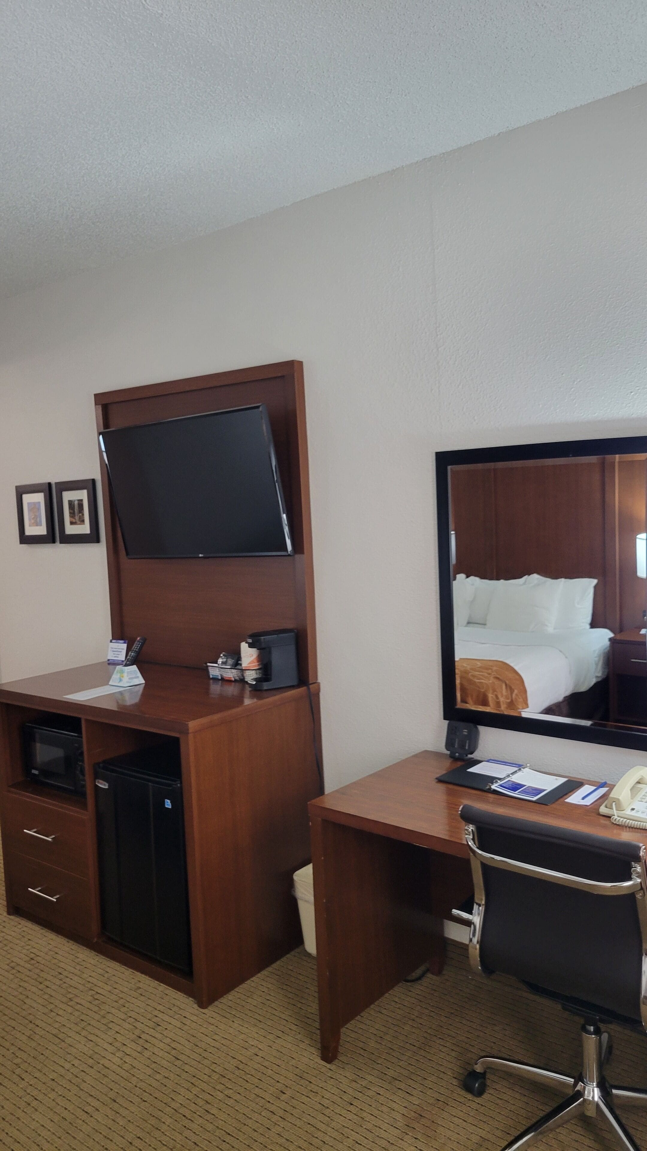A hotel room with a wooden desk, a flat-screen TV, a mini-fridge, a microwave, a chair, a bed with white linens, and a large mirror.
