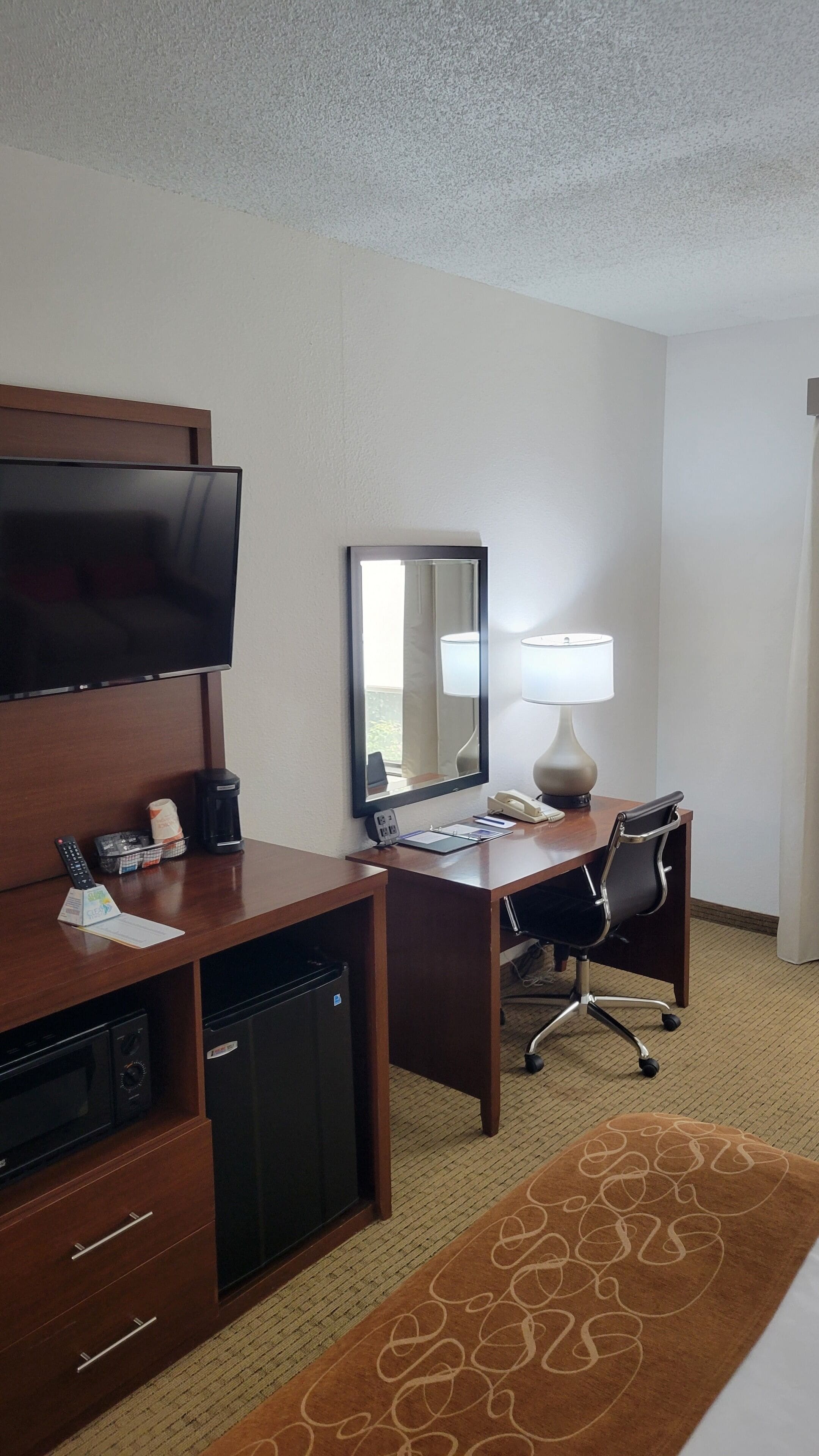 Habitación de hotel con televisión de pantalla plana, un escritorio con silla, una lámpara, cafetera y refrigerador pequeño.