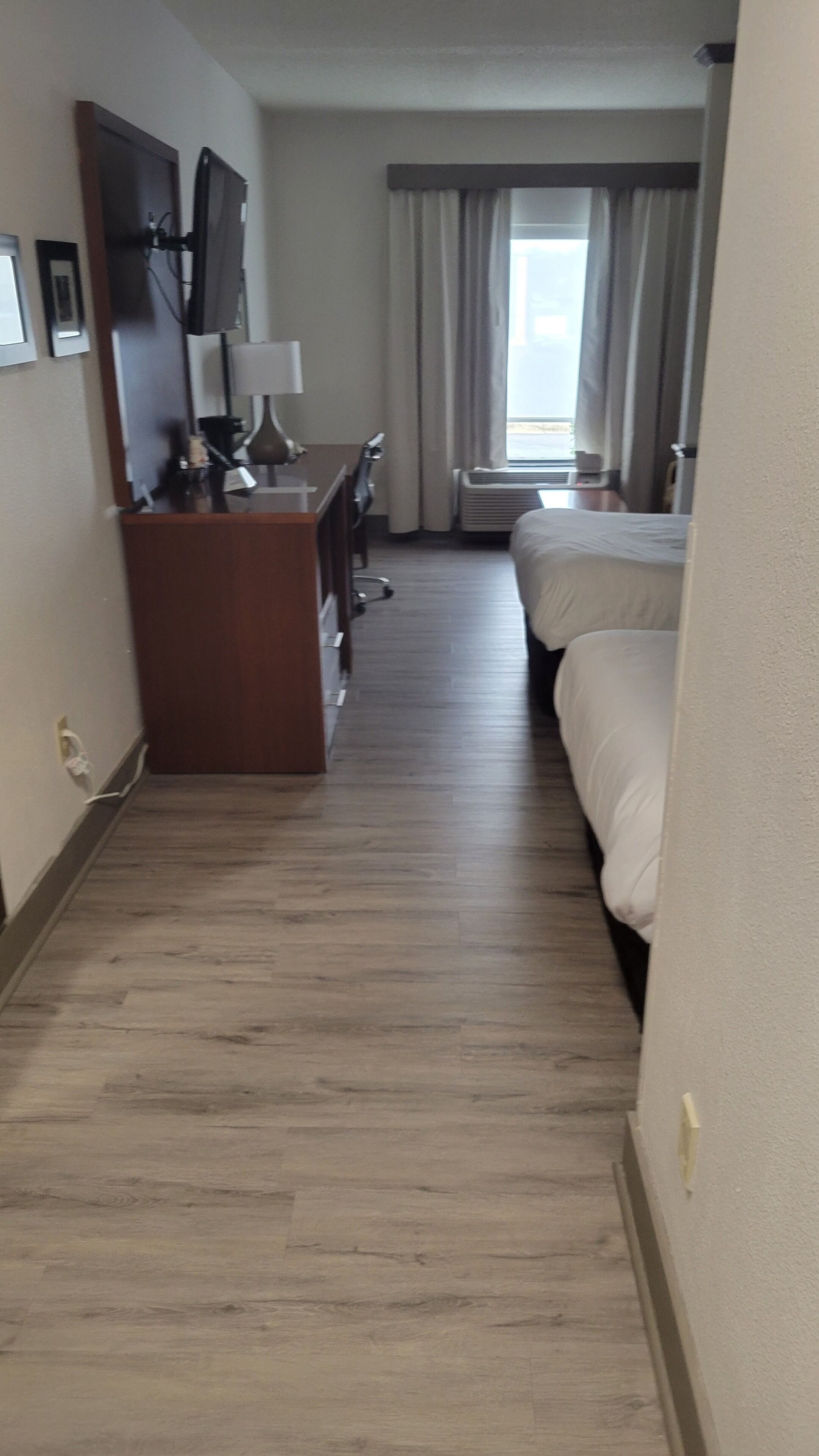 A hotel room with a wooden floor, a desk with a lamp, a bed, and a window with curtains.