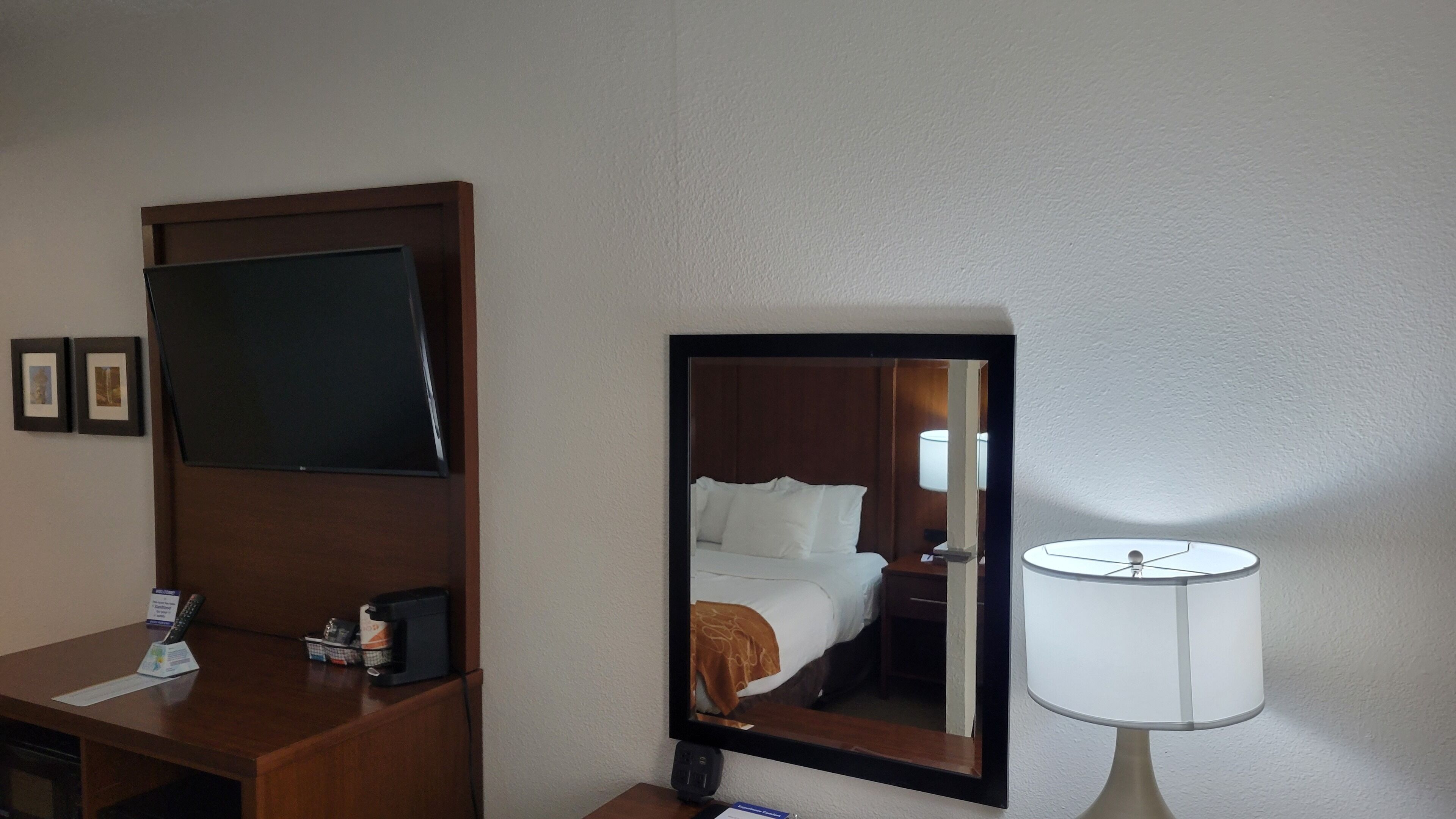 A hotel room with a flat-screen TV mounted on the wall, a bedside table with a lamp, and a bed with a brown blanket.