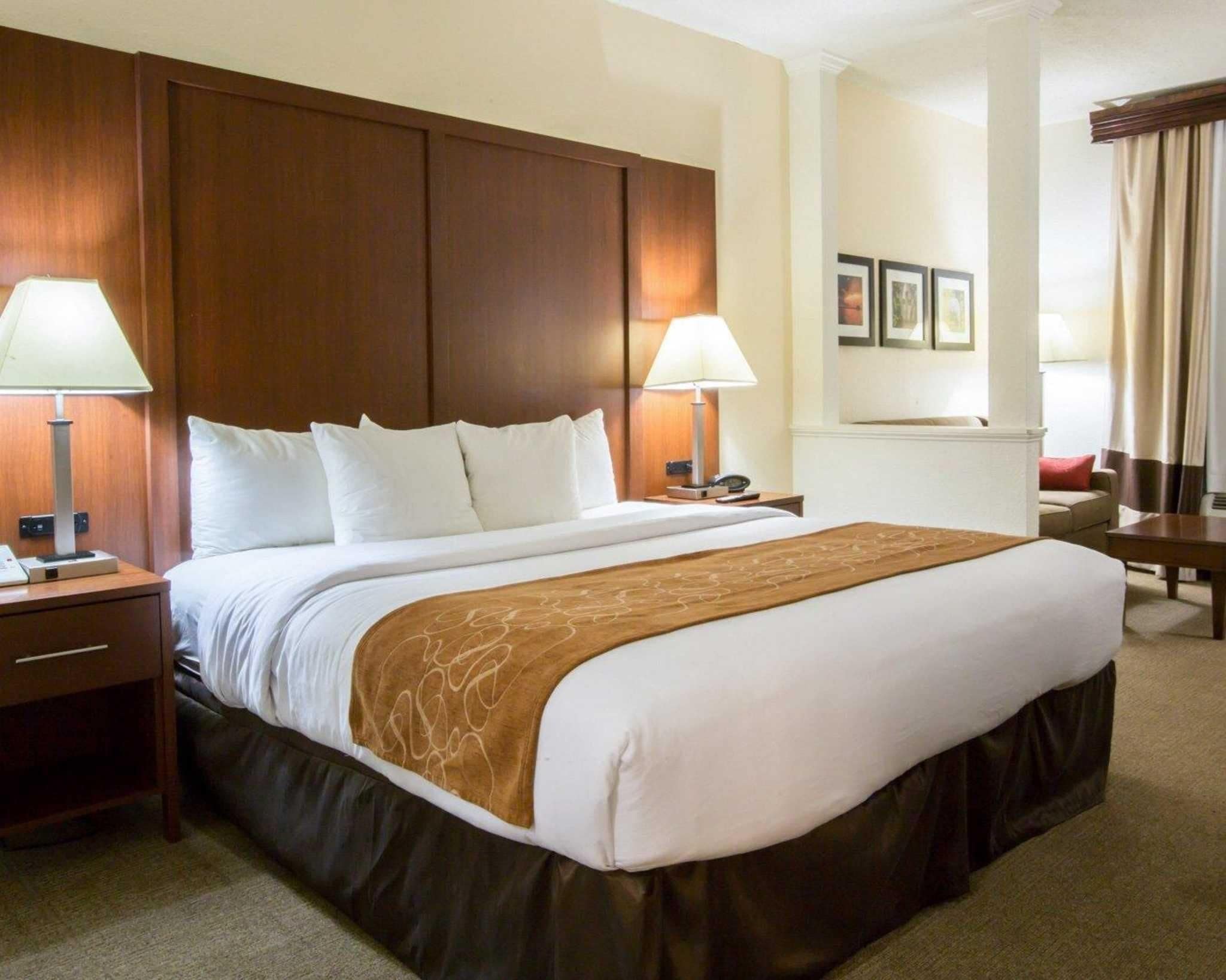 Quarto de hotel com uma cama grande, duas mesas de cabeceira, duas luminárias, uma cabeceira de madeira, um cobertor marrom, um travesseiro vermelho, uma mesinha e uma cadeira.